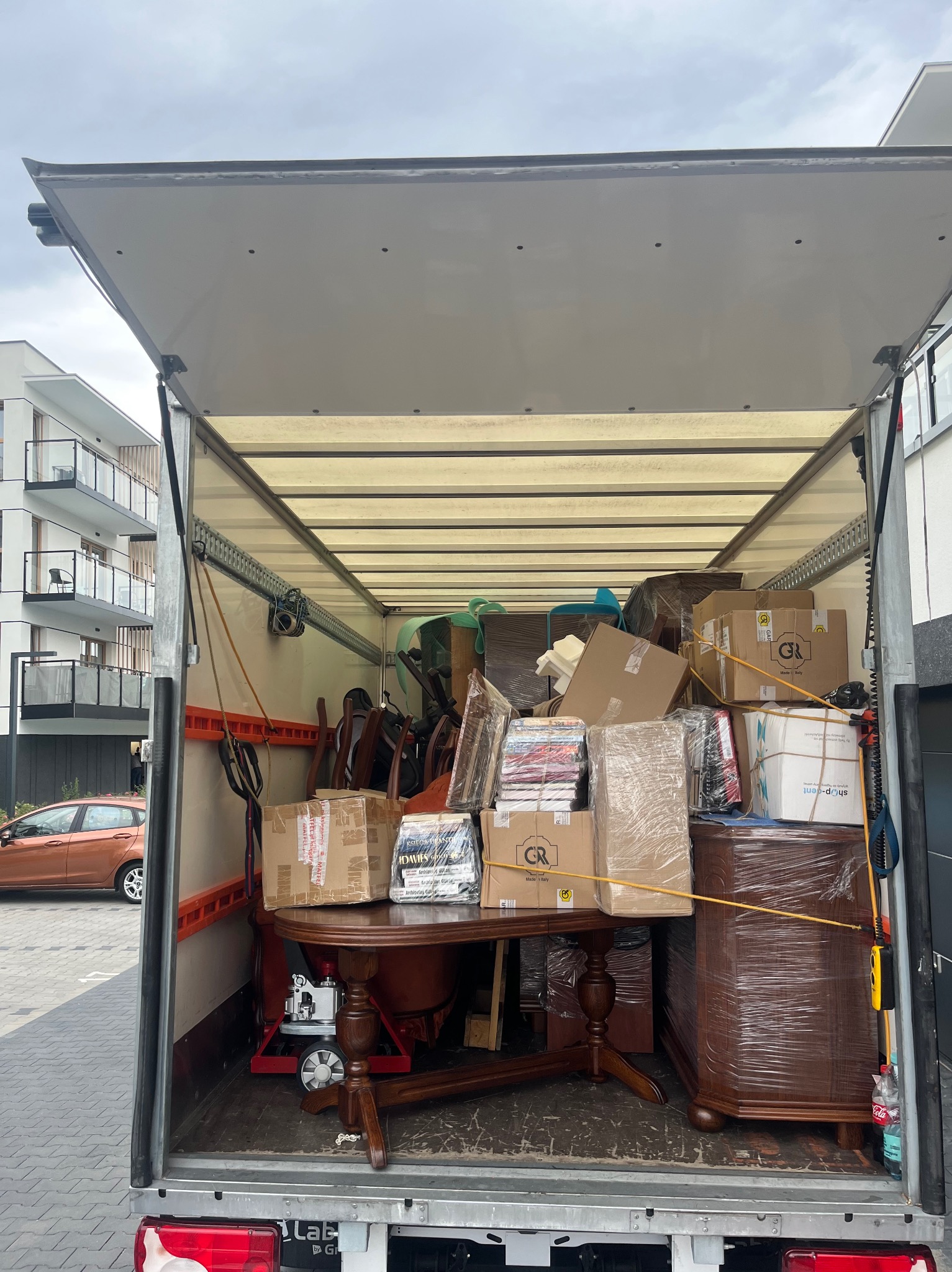 Wnętrze busa załadowane meblami i kartonami, zabezpieczonymi do transportu. Widoczny stół z krzesłami, komoda owinięta folią. W tle budynek mieszkalny.