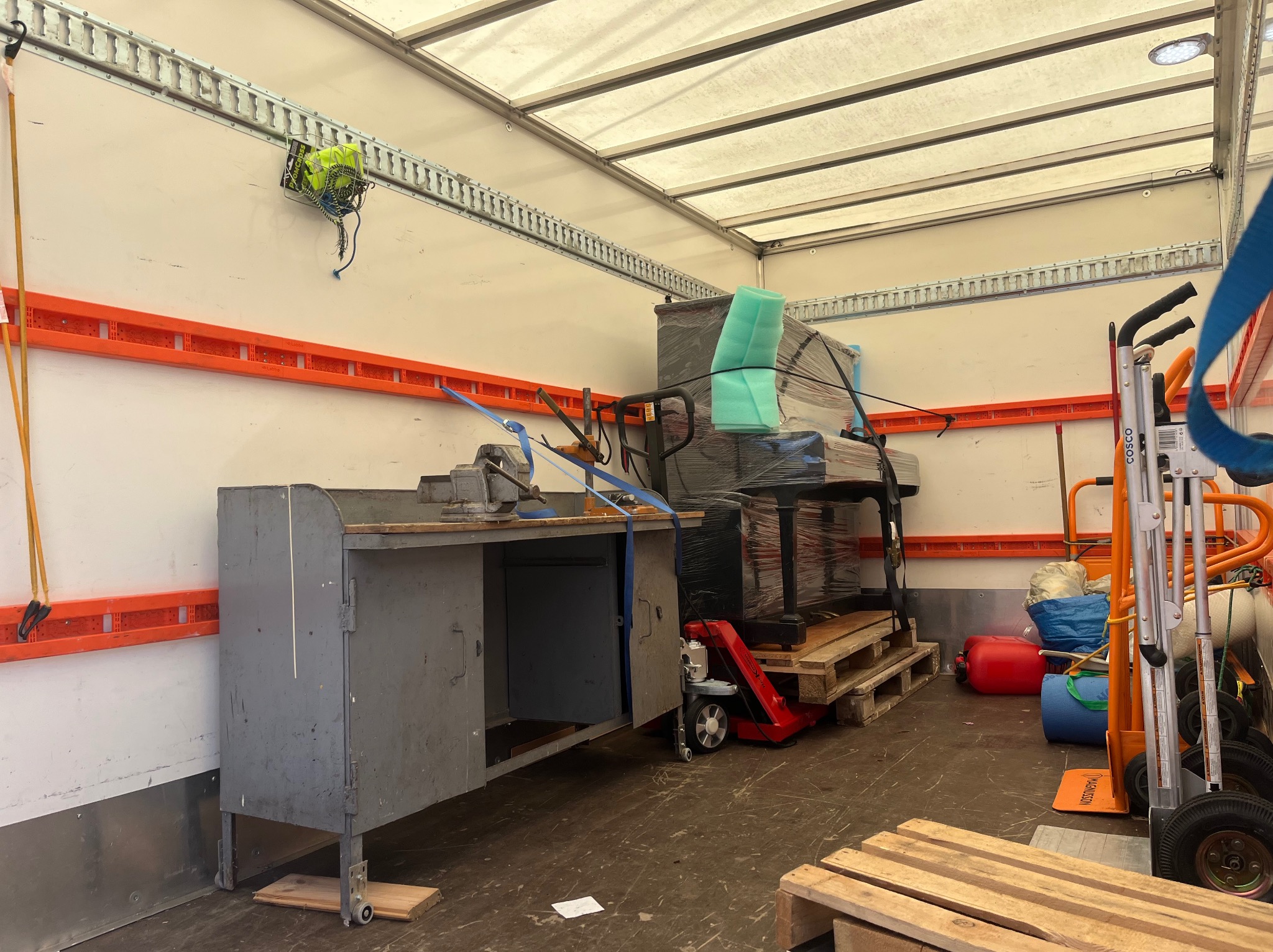 Wnętrze furgonetki załadowanej meblami i sprzętem zabezpieczonym folią, z wózkiem transportowym i stołem warsztatowym z imadłem po lewej stronie.