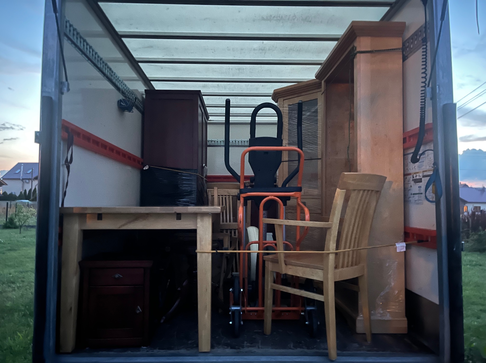 Wnętrze furgonetki załadowanej meblami i sprzętem do ćwiczeń, przygotowanymi do przeprowadzki. Widoczny stół, krzesła, komoda i orbitrek zabezpieczone do transportu.