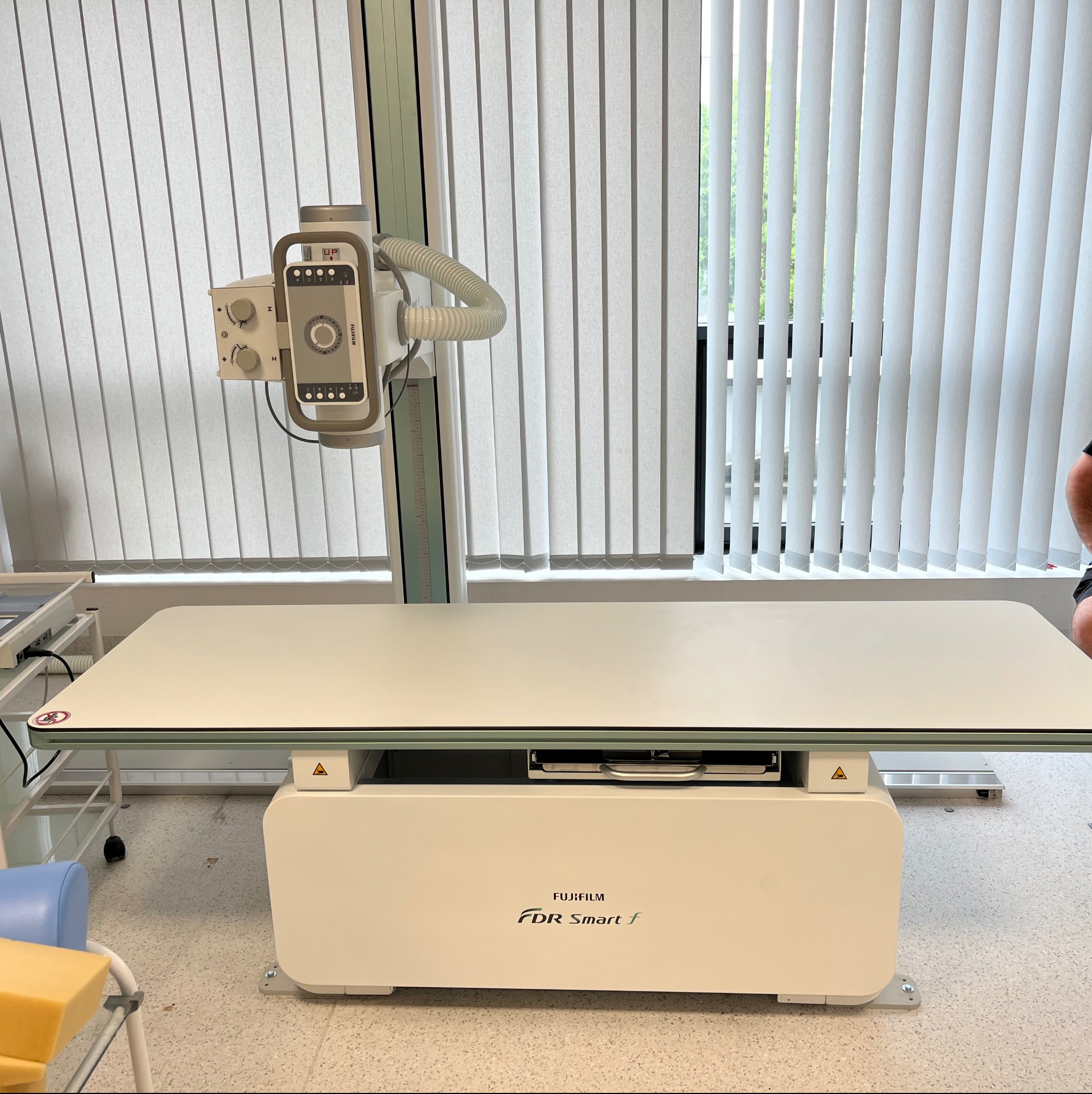 Zdjęcie przedstawia aparat rentgenowski Fujifilm FDR Smart f w jasnym pomieszczeniu z żaluzjami. Widoczny stół do badań i panel sterowania.