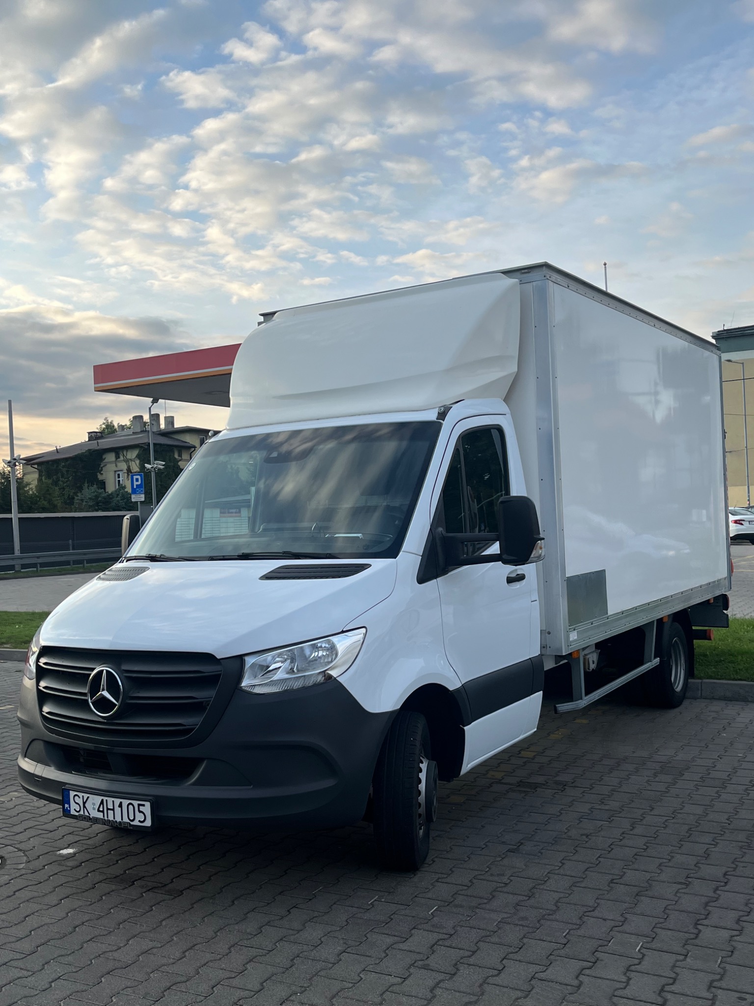Biały samochód dostawczy Mercedes-Benz z zabudową kontenerową na parkingu z kostki brukowej, widoczny znak stacji paliw w tle.