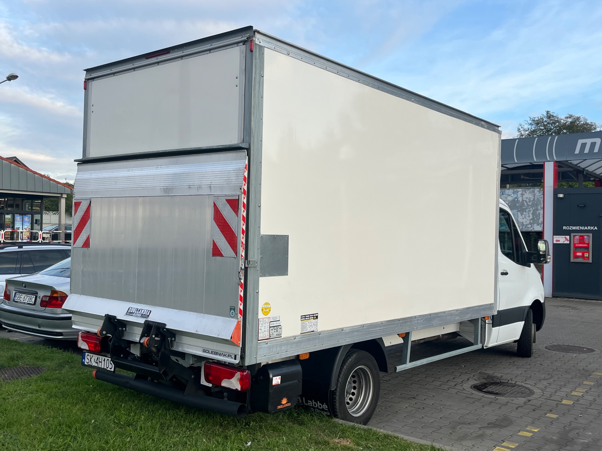 Biały samochód dostawczy z windą załadowczą, zaparkowany na zewnątrz, widziany z tyłu pod kątem. Widoczne oznaczenia i tablica rejestracyjna.