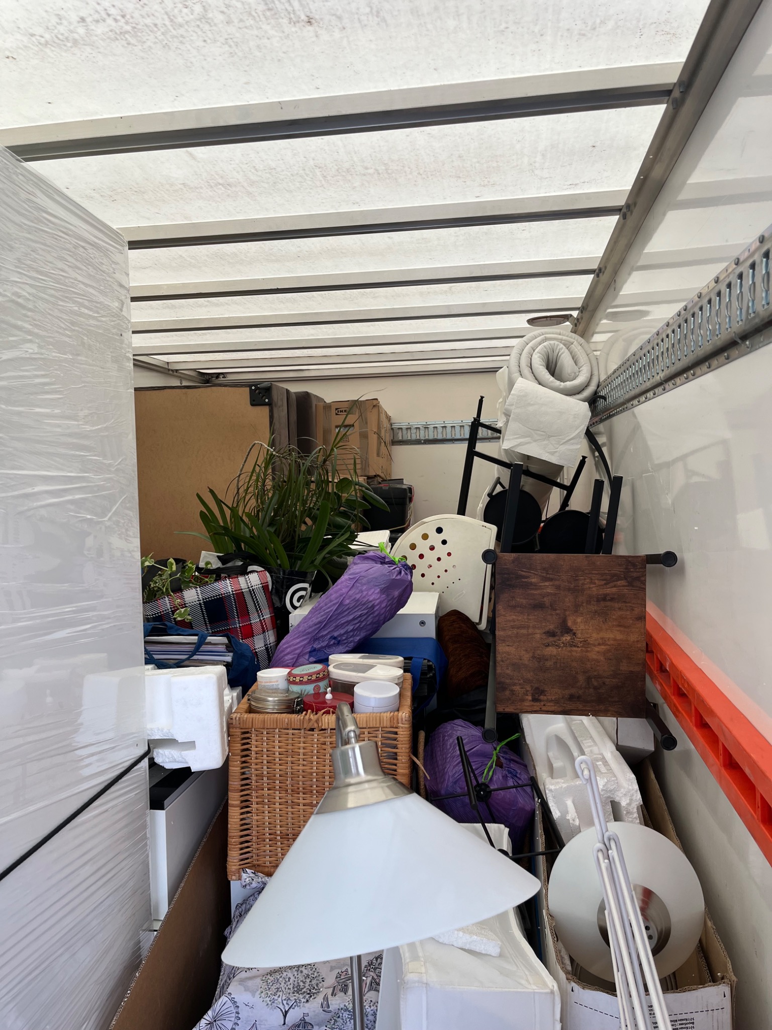 Wnętrze furgonetki wypełnionej różnorodnymi przedmiotami, w tym meblami, roślinami i pudełkami, przygotowanymi do transportu. Widoczny fragment lampy i krzesła.