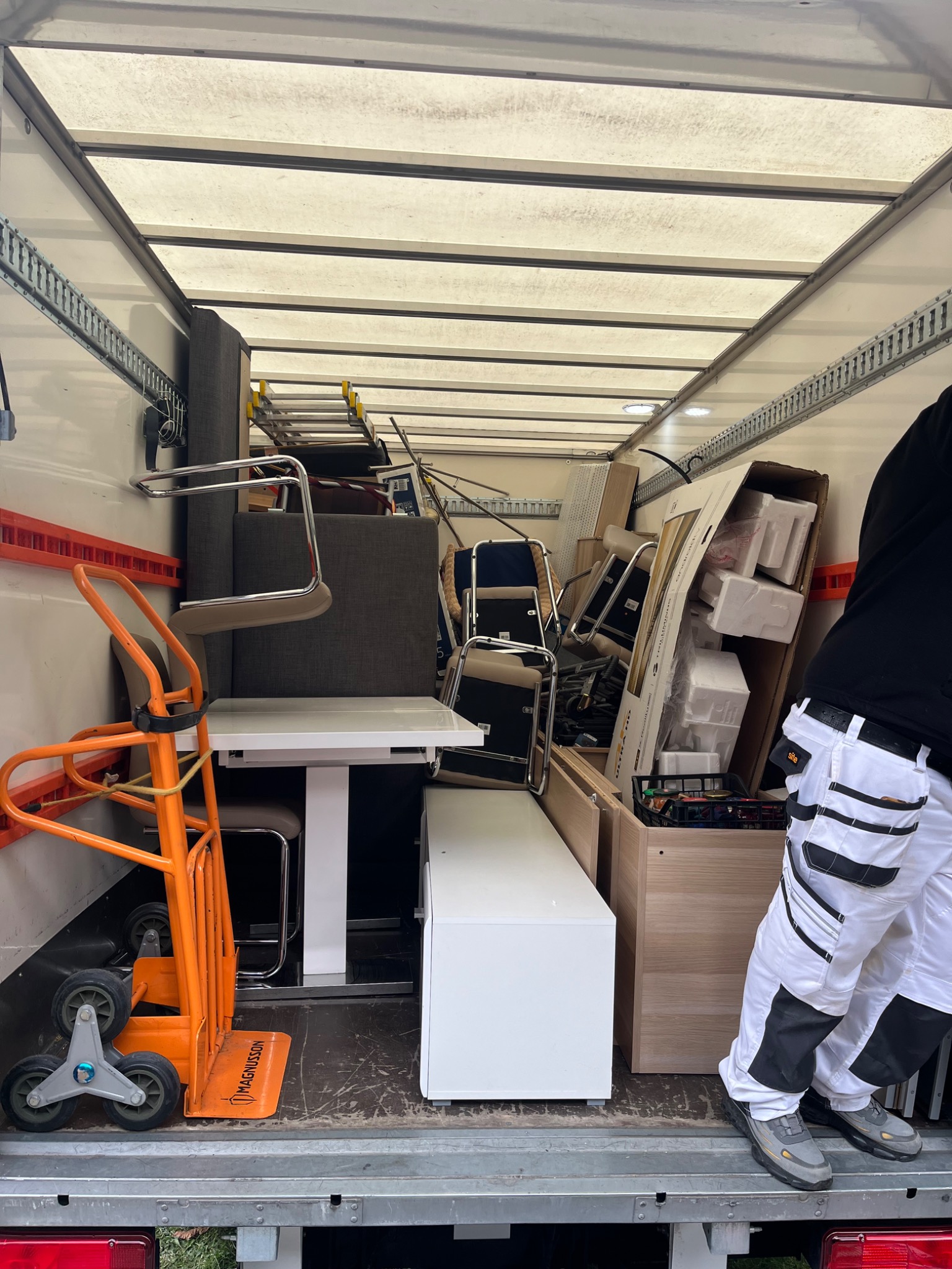 Wnętrze furgonetki wypełnionej meblami i sprzętami biurowymi podczas przeprowadzki. Widoczny wózek transportowy i fragment osoby w stroju roboczym.