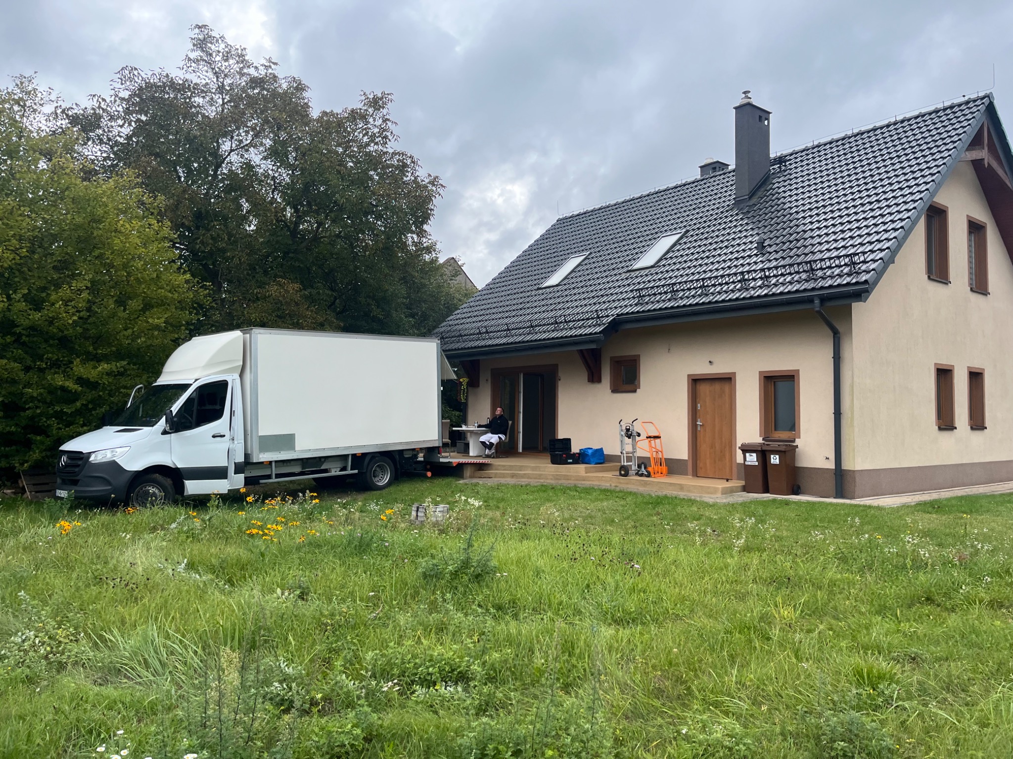 Przeprowadzka: biały samochód dostawczy zaparkowany przed domem z osobą siedzącą na platformie załadunkowej. W tle widoczny trawnik i elementy wyposażenia.