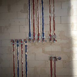 RK Instal Roman Naholiuk - Instalacja rur wodnych (czerwone i niebieskie) w ścianie z białego pustaka, zabezpieczonych pianką montażową. Widoczne zawory i manometry.