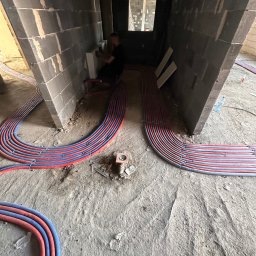 RK Instal Roman Naholiuk - Instalacja ogrzewania podłogowego w surowym wnętrzu, widoczne rury grzewcze ułożone na posadzce, pracownik montuje grzejnik w tle. Instalacje grzewcze w trakcie realizacji.