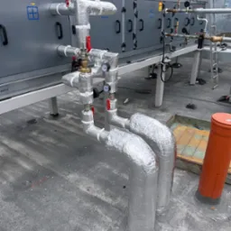 Izolowane rury instalacji przemysłowej na dachu budynku, z zaworami i manometrami, na tle szarej obudowy urządzenia klimatyzacyjnego.