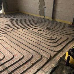 RK Instal Roman Naholiuk - Ułożone rury ogrzewania podłogowego na folii z czerwoną siatką, widoczne elementy instalacji i fragment sylwetki osoby w rogu, surowy stan.