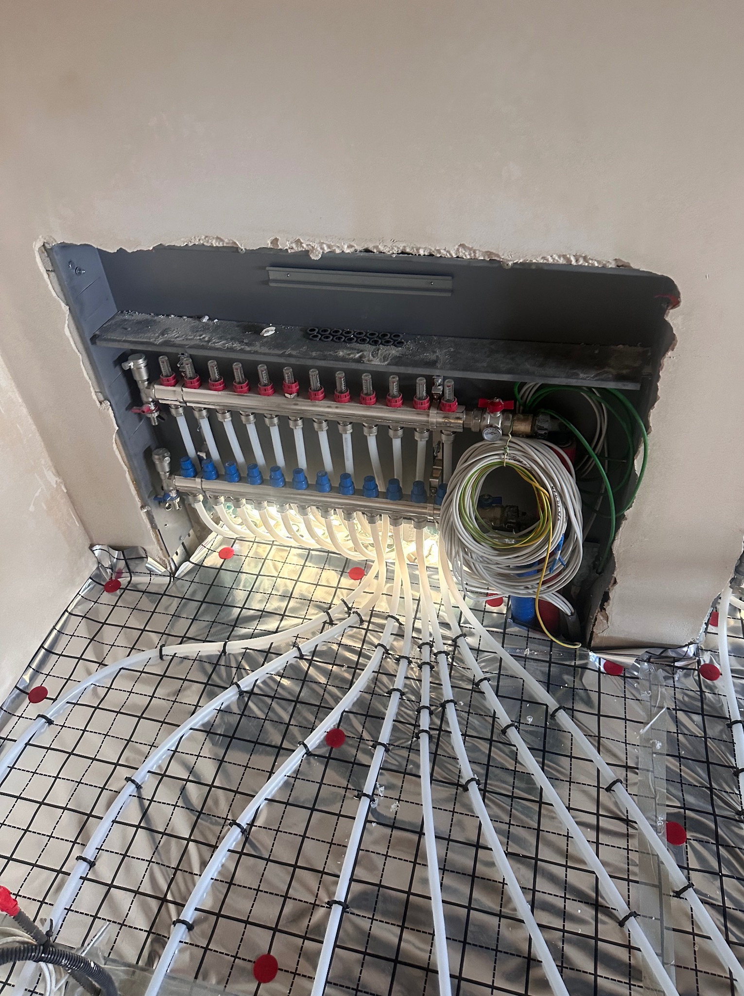 Rozdzielacz ogrzewania podłogowego z rurami ułożonymi na izolacji, widok z góry, instalacja w trakcie montażu, przewody elektryczne obok rozdzielacza.