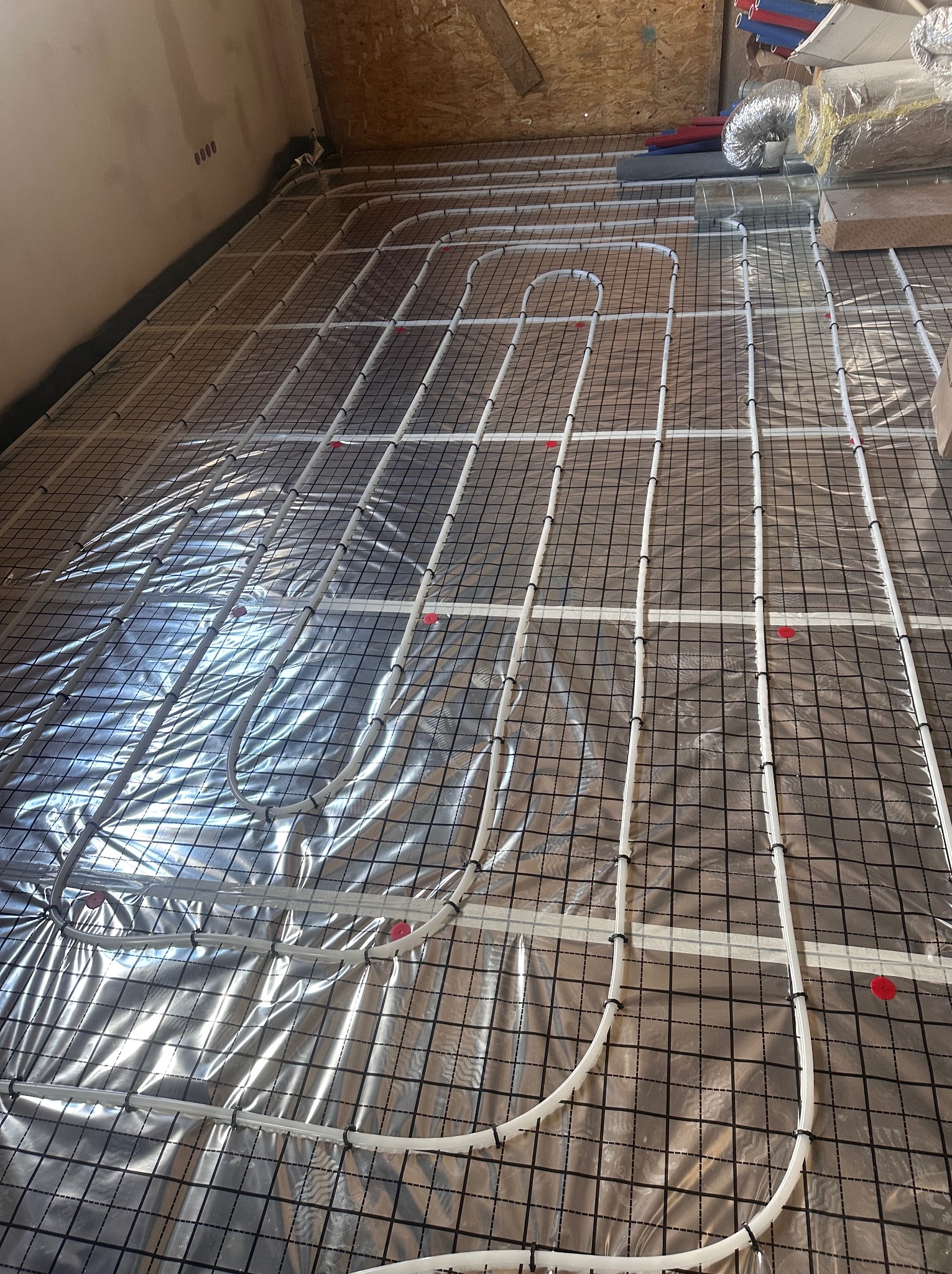 Białe rury ogrzewania podłogowego, zamocowane na folii izolacyjnej z rastrem, w surowym wnętrzu. Elementy instalacji grzewczej widoczne w tle.