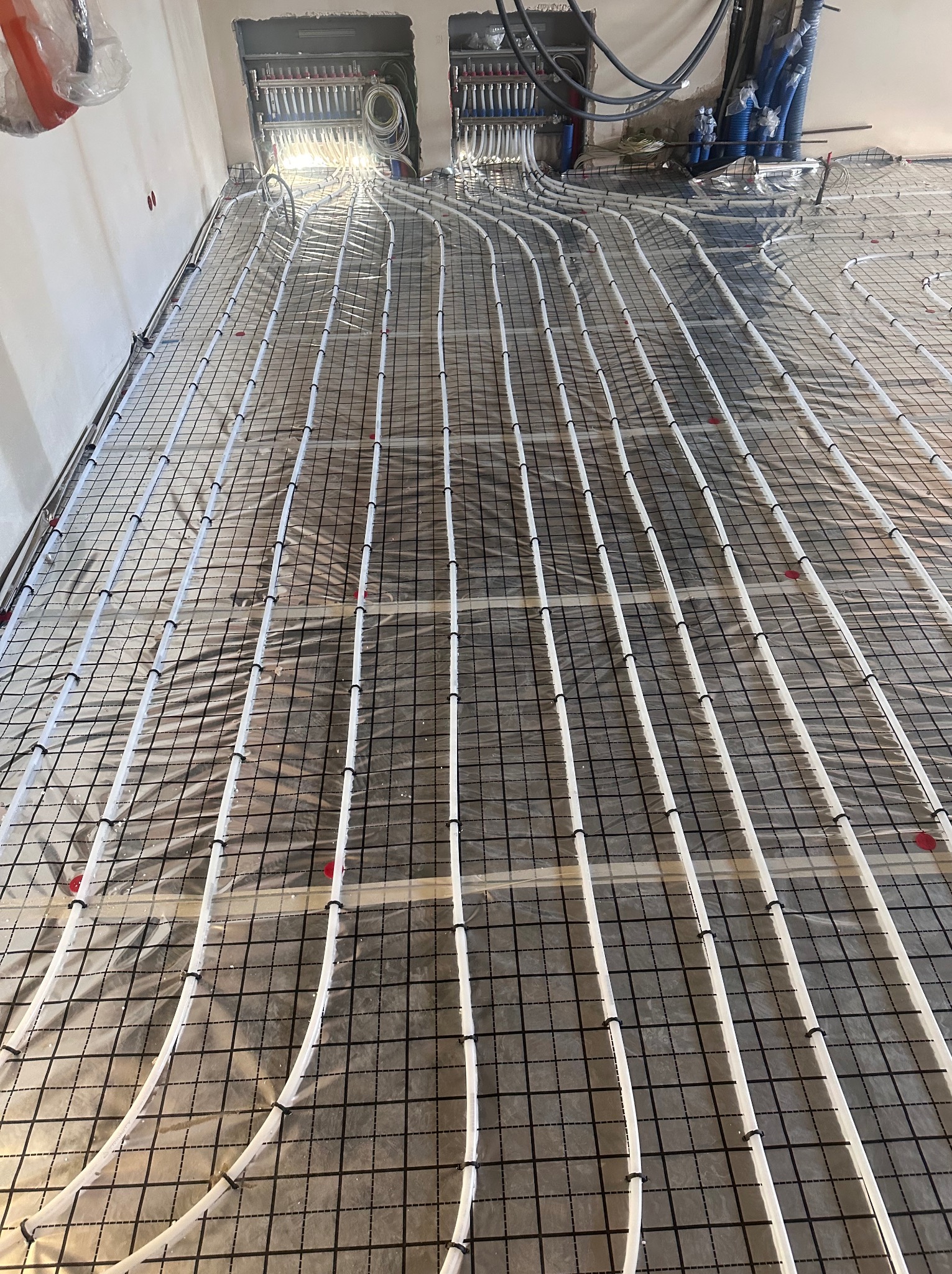 Białe rury ogrzewania podłogowego, zamocowane do czarnej siatki, rozchodzą się promieniście od rozdzielaczy w surowym stanie deweloperskim.