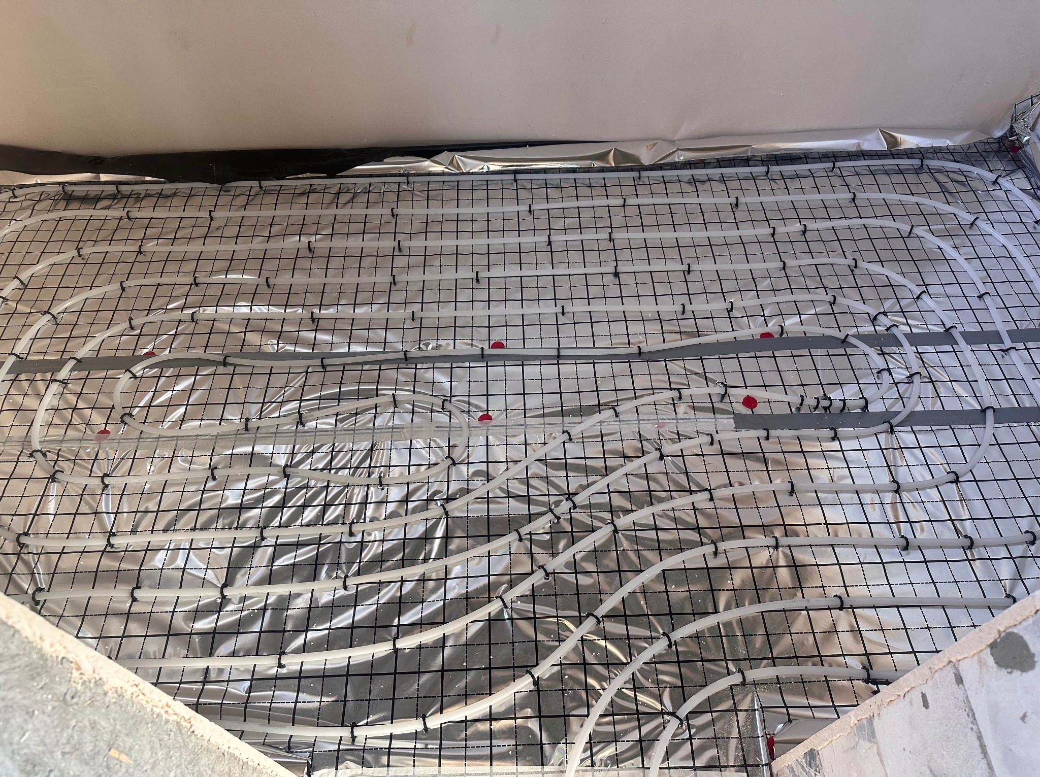 Białe rury ogrzewania podłogowego ułożone na srebrnej folii, przymocowane do czarnej siatki. Widok z góry, instalacja w trakcie realizacji.