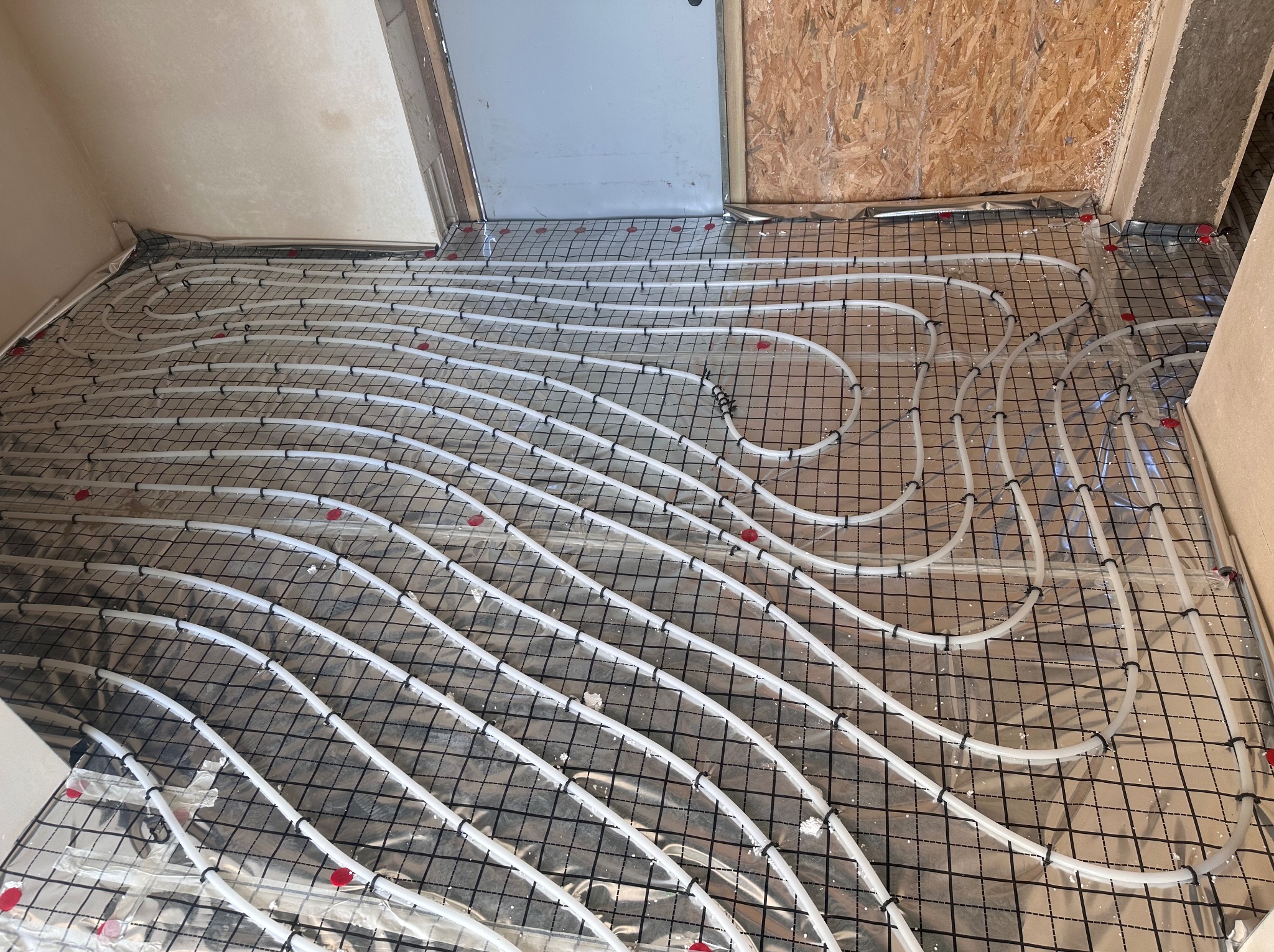 Białe rury ogrzewania podłogowego ułożone na czarnej siatce, na podłożu z folii aluminiowej. Widok na fragment podłogi w trakcie montażu systemu grzewczego.