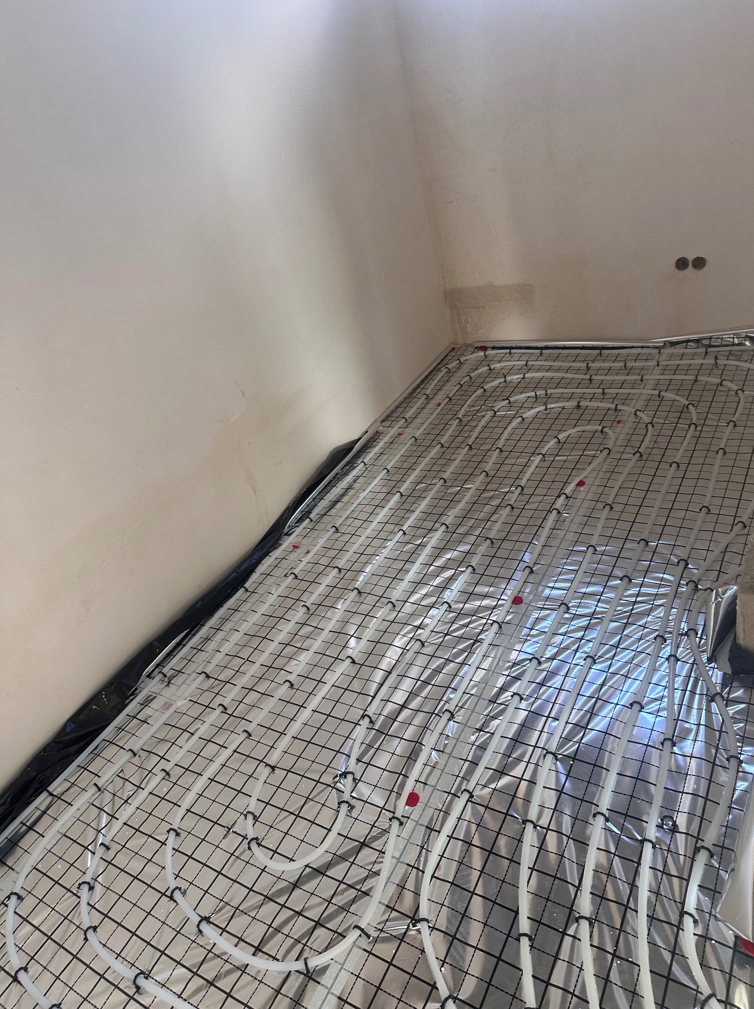 Białe rury ogrzewania podłogowego ułożone na srebrnej folii z czarną siatką w surowym wnętrzu. Widok z góry, blisko ściany z widocznymi śladami po instalacjach.