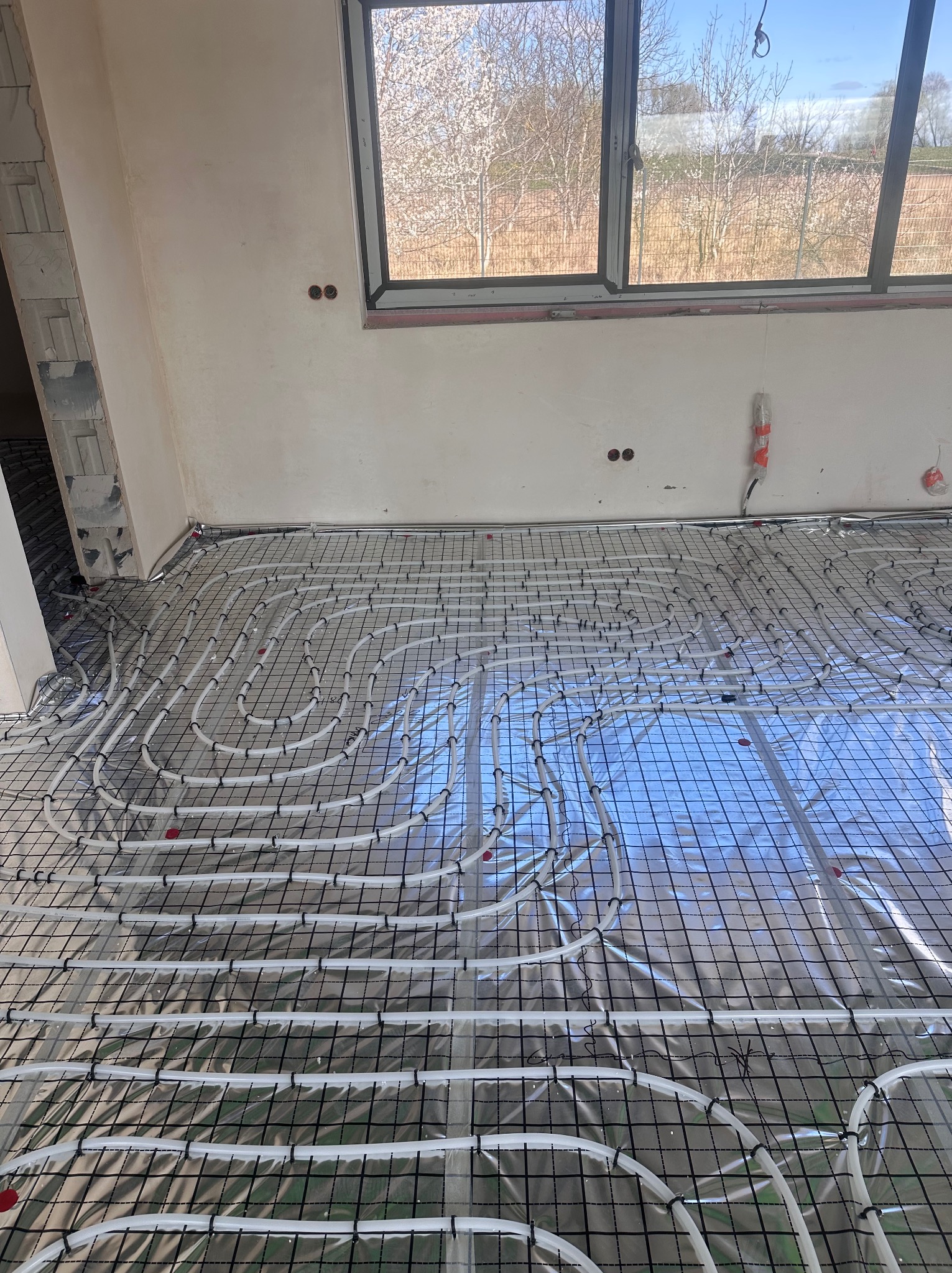 Białe rury ogrzewania podłogowego ułożone na folii i siatce w jasnym pomieszczeniu z oknem. Widoczne elementy instalacji, etap prac przed wylaniem posadzki.