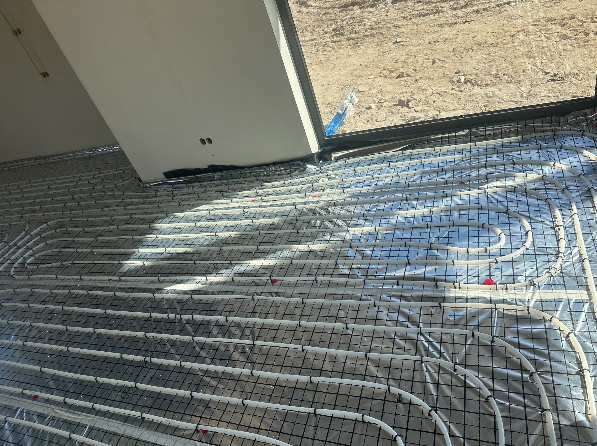 Białe rury ogrzewania podłogowego ułożone na folii odblaskowej i siatce, widok z góry, część rur pod oknem. Instalacja w trakcie realizacji.