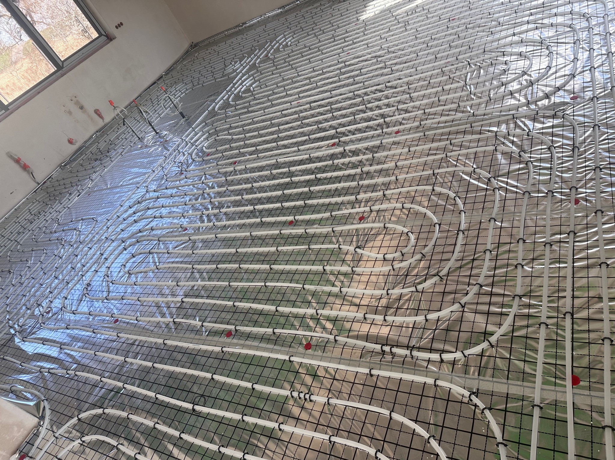 Białe rury ogrzewania podłogowego ułożone na srebrnej folii z czarną siatką, widoczne z góry, przed wylaniem posadzki. Fragment okna w tle.