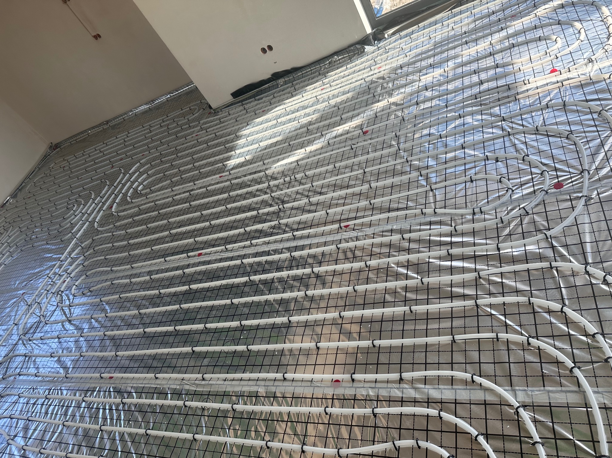 Białe rury ogrzewania podłogowego ułożone na srebrnej folii z czarną siatką, widok z góry, jasne wnętrze z oknem w tle.