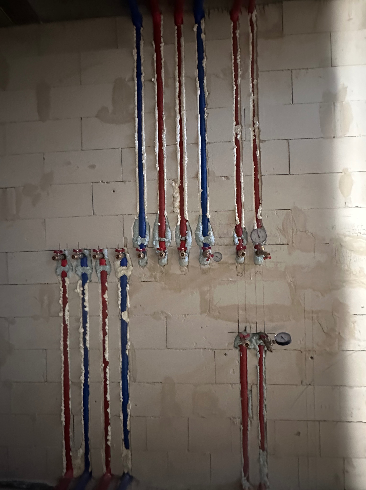 Instalacja rur wodnych (czerwone i niebieskie) w ścianie z białego pustaka, zabezpieczonych pianką montażową. Widoczne zawory i manometry.