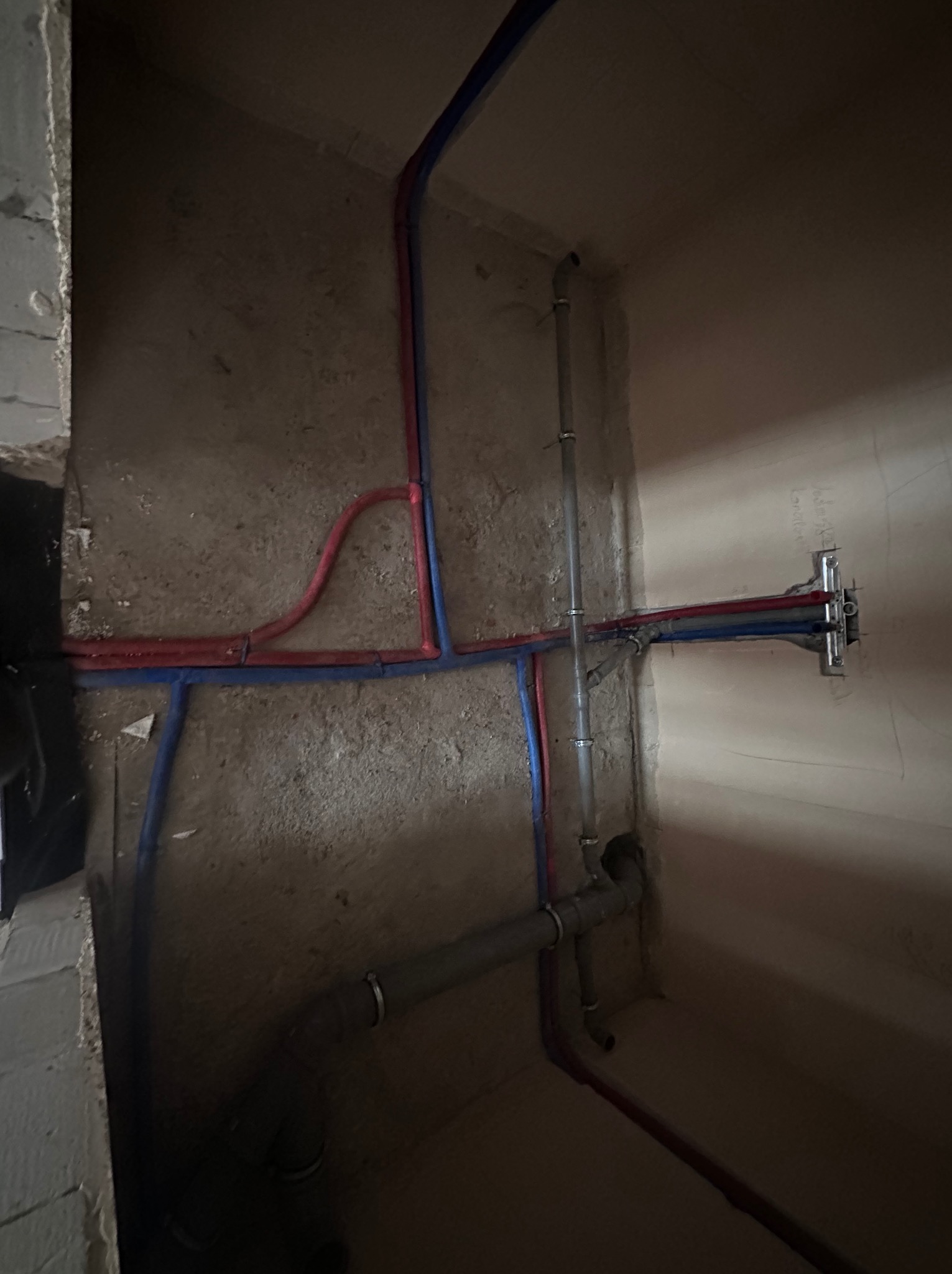 Surowe ściany z instalacją hydrauliczną: rury miedziane i plastikowe w kolorze czerwonym i niebieskim, pionowe rury kanalizacyjne z uchwytami, widok z dołu.