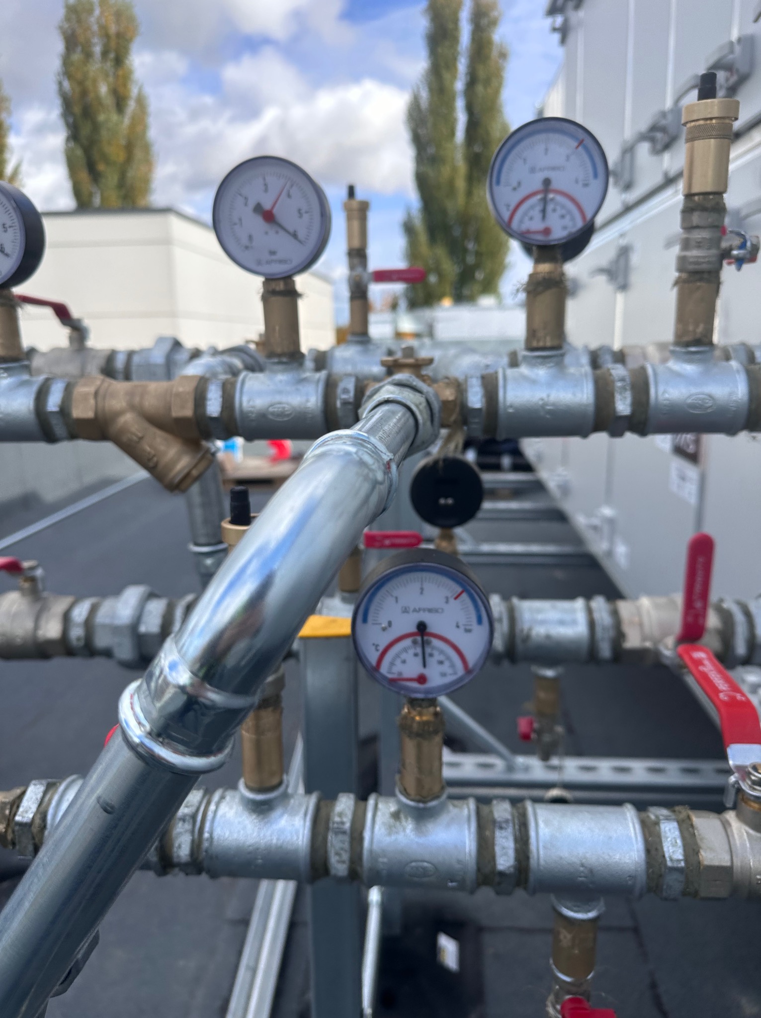 Złożona instalacja hydrauliczna z manometrami na zewnątrz budynku, widoczne liczne rury, zawory i filtry. Tło stanowią drzewa i błękitne niebo.