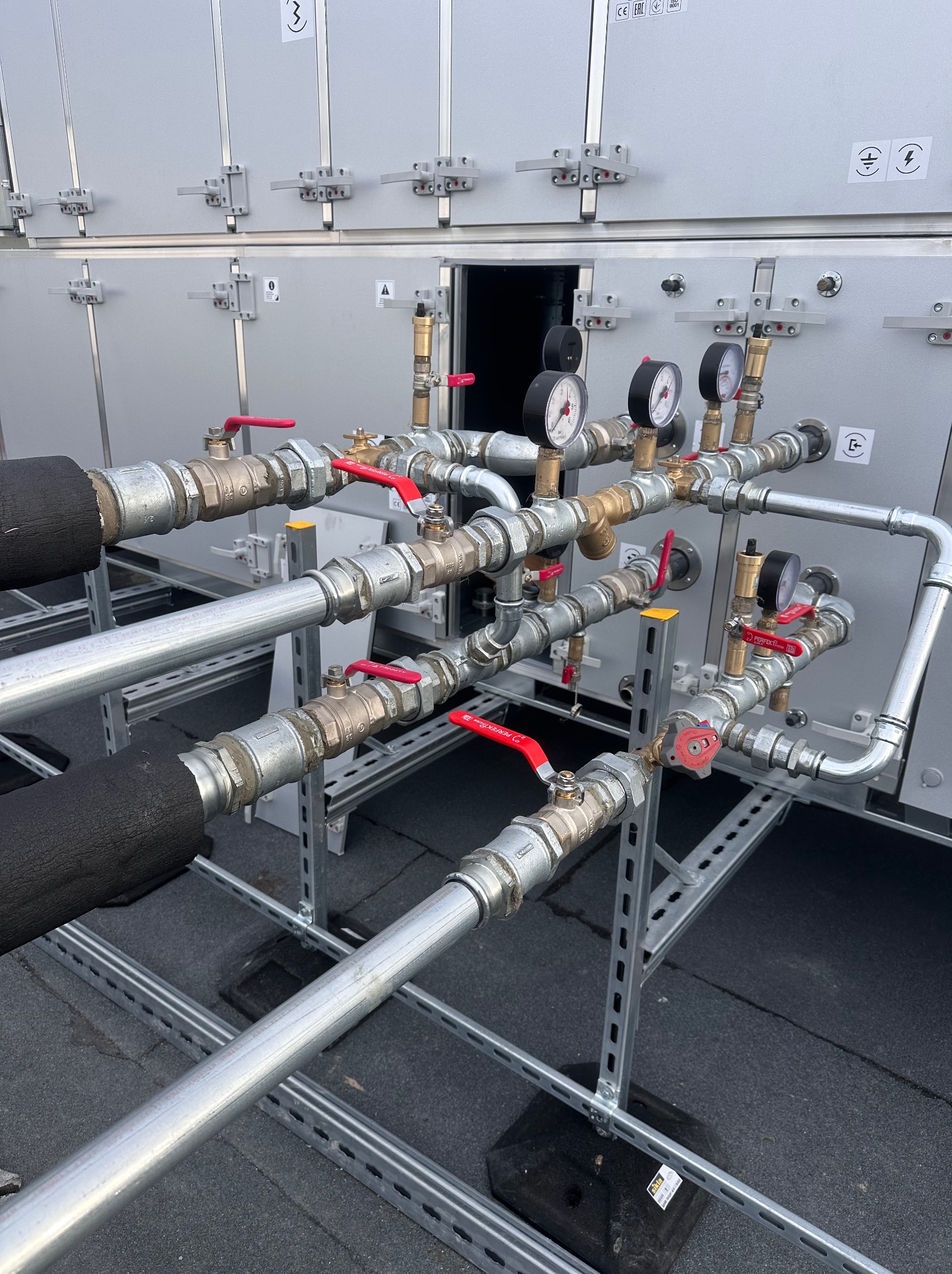 Złożona instalacja rurowa z zaworami i manometrami na metalowej konstrukcji wsporczej, na tle szarej obudowy. Widoczne czarne izolacje termiczne na rurach.