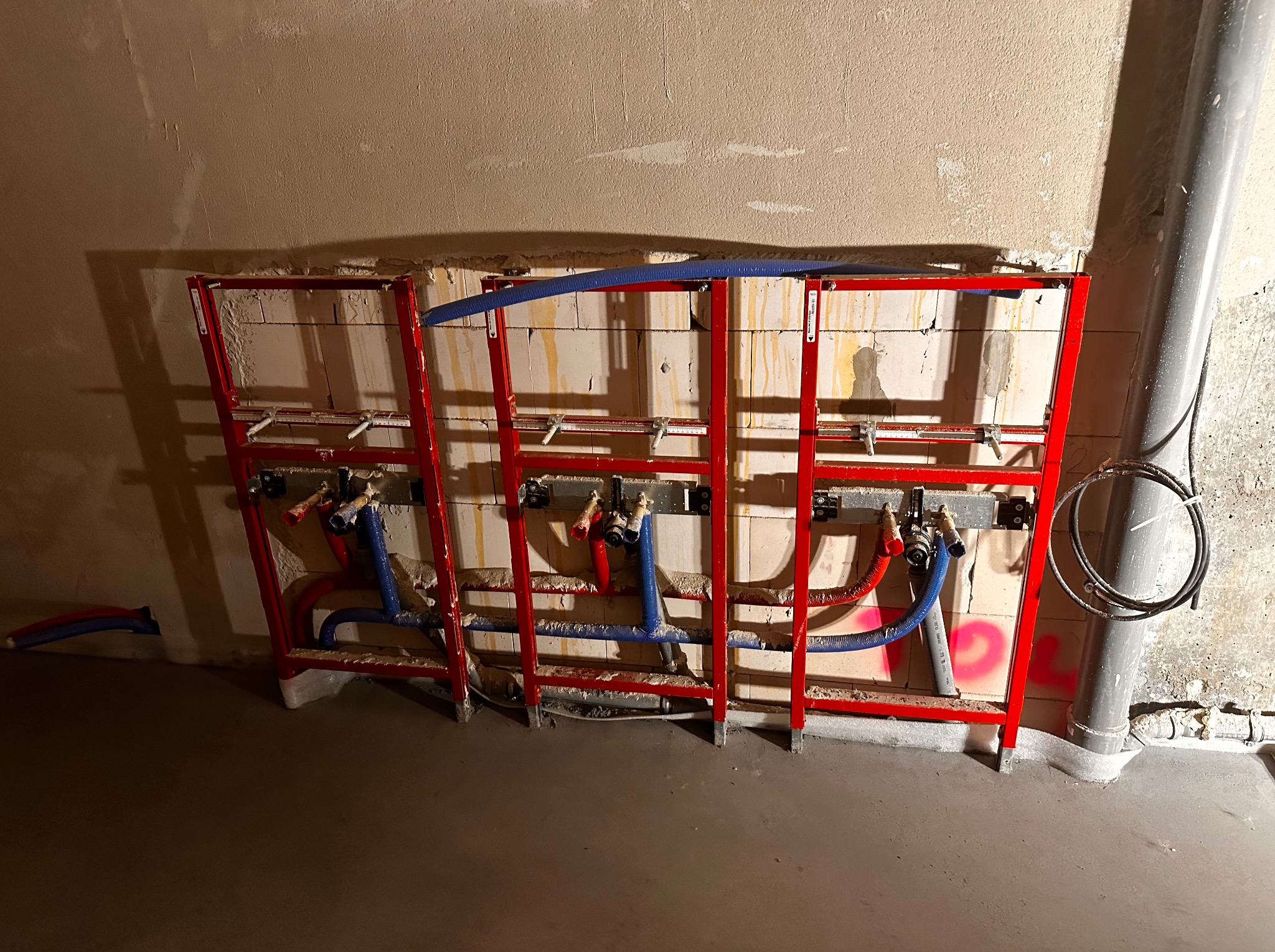 Czerwona rama montażowa z niebieskimi i czerwonymi rurami na tle ściany z pustaków i szarej rury kanalizacyjnej. Widok na instalację wodną przed zabudową.