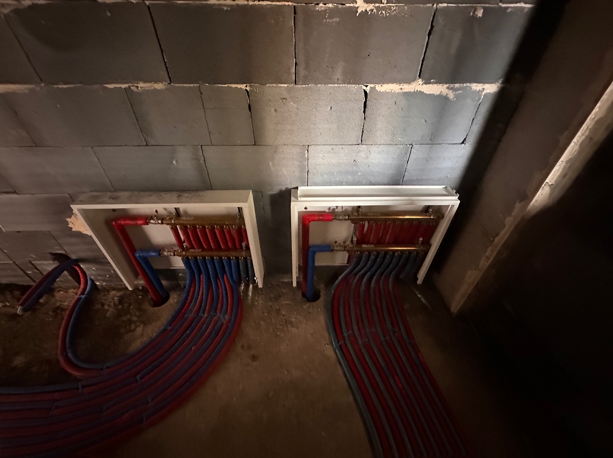 Rozdzielacze ogrzewania podłogowego z rurami w kolorach czerwonym i niebieskim, zamontowane na szarej ścianie z pustaków. Widok z góry, w trakcie instalacji.