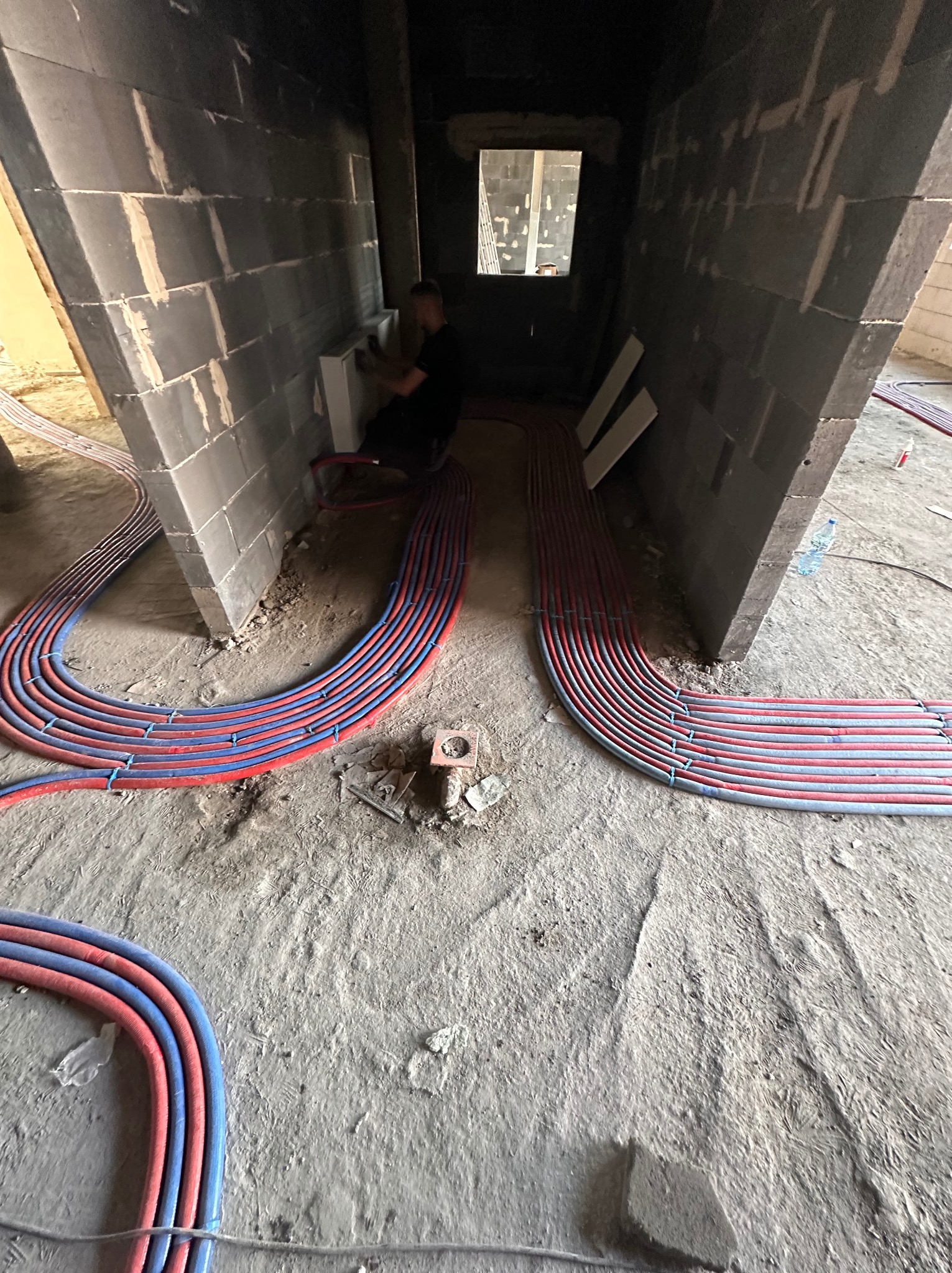 Instalacja ogrzewania podłogowego w surowym wnętrzu, widoczne rury grzewcze ułożone na posadzce, pracownik montuje grzejnik w tle. Instalacje grzewcze w trakcie realizacji.