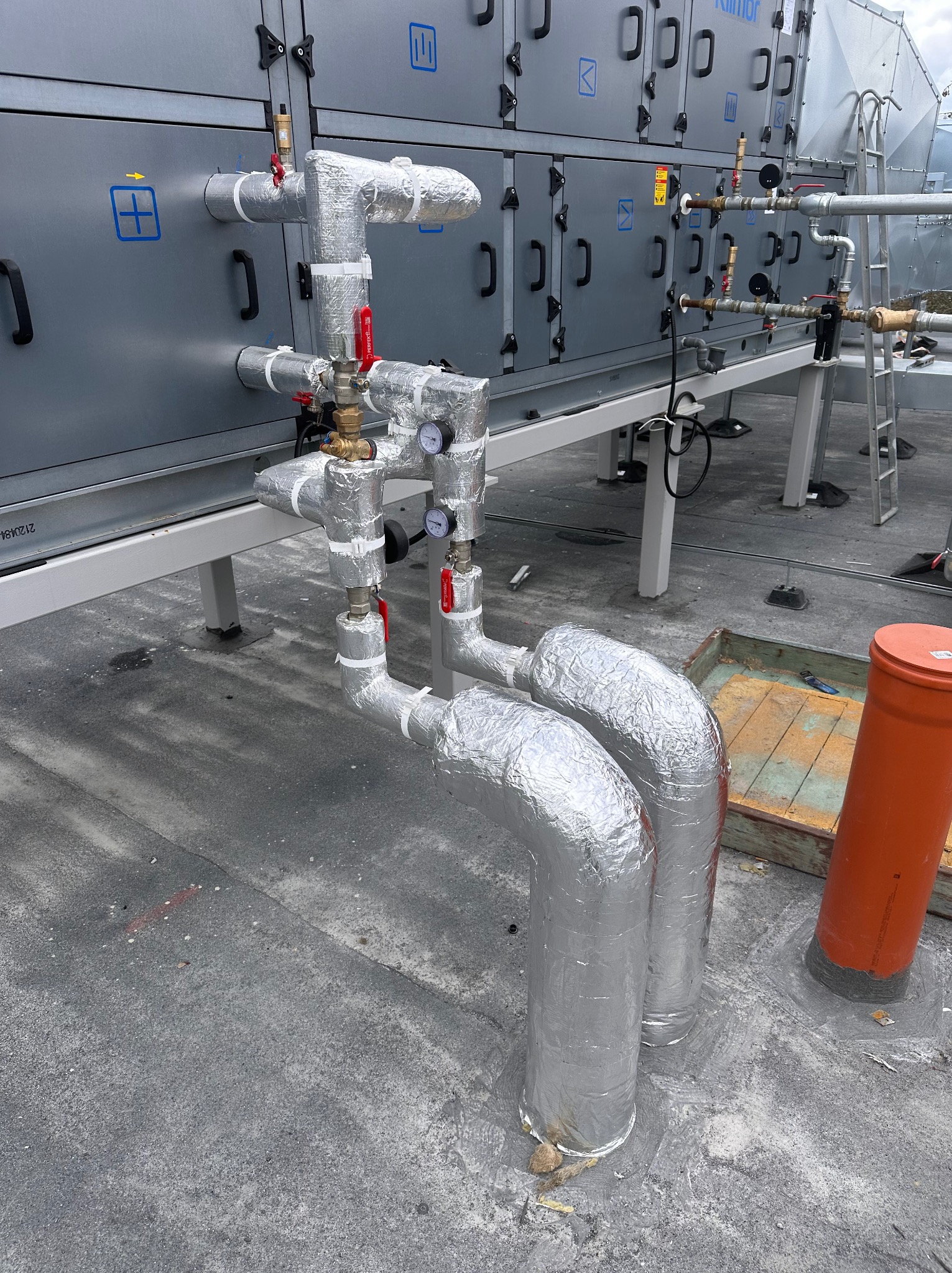 Izolowane rury instalacji przemysłowej na dachu budynku, z zaworami i manometrami, na tle szarej obudowy urządzenia klimatyzacyjnego.