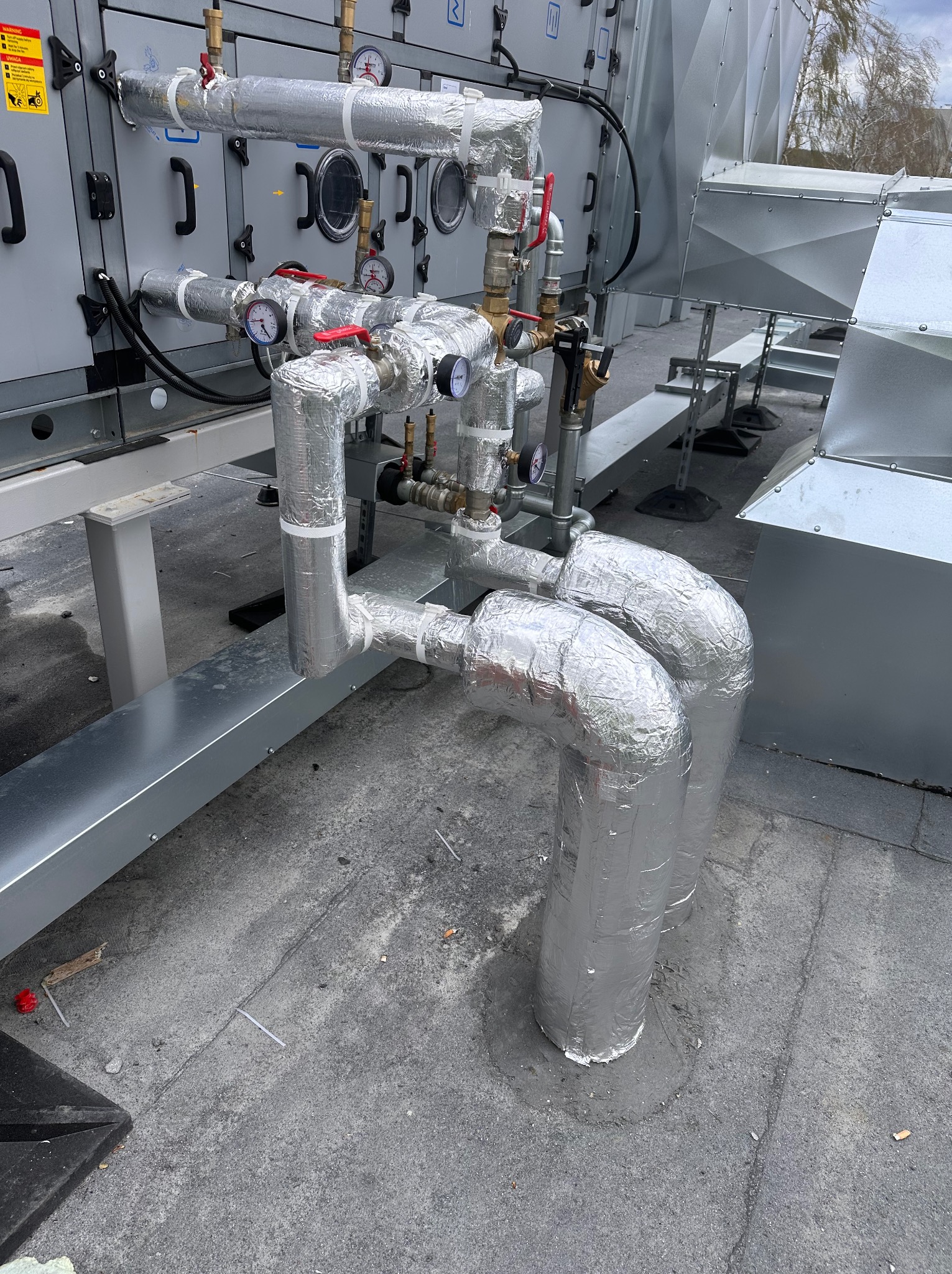 Izolowane rury instalacji przemysłowej na dachu z zaworami, manometrami i elementami wentylacji. Srebrna folia izolacyjna z taśmą. Szara betonowa powierzchnia dachu.