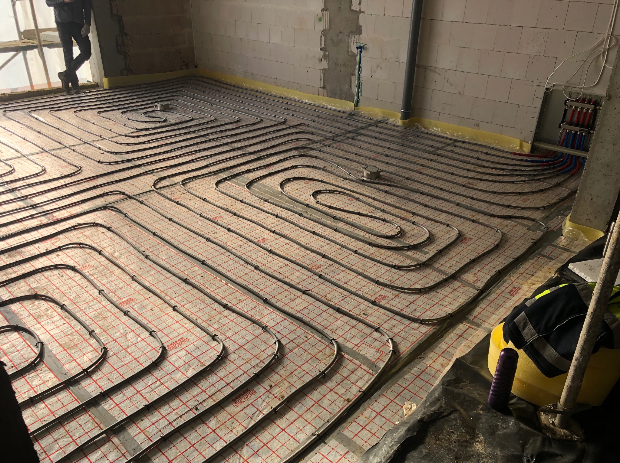 Ułożone rury ogrzewania podłogowego na folii z czerwoną siatką, widoczne elementy instalacji i fragment sylwetki osoby w rogu, surowy stan.