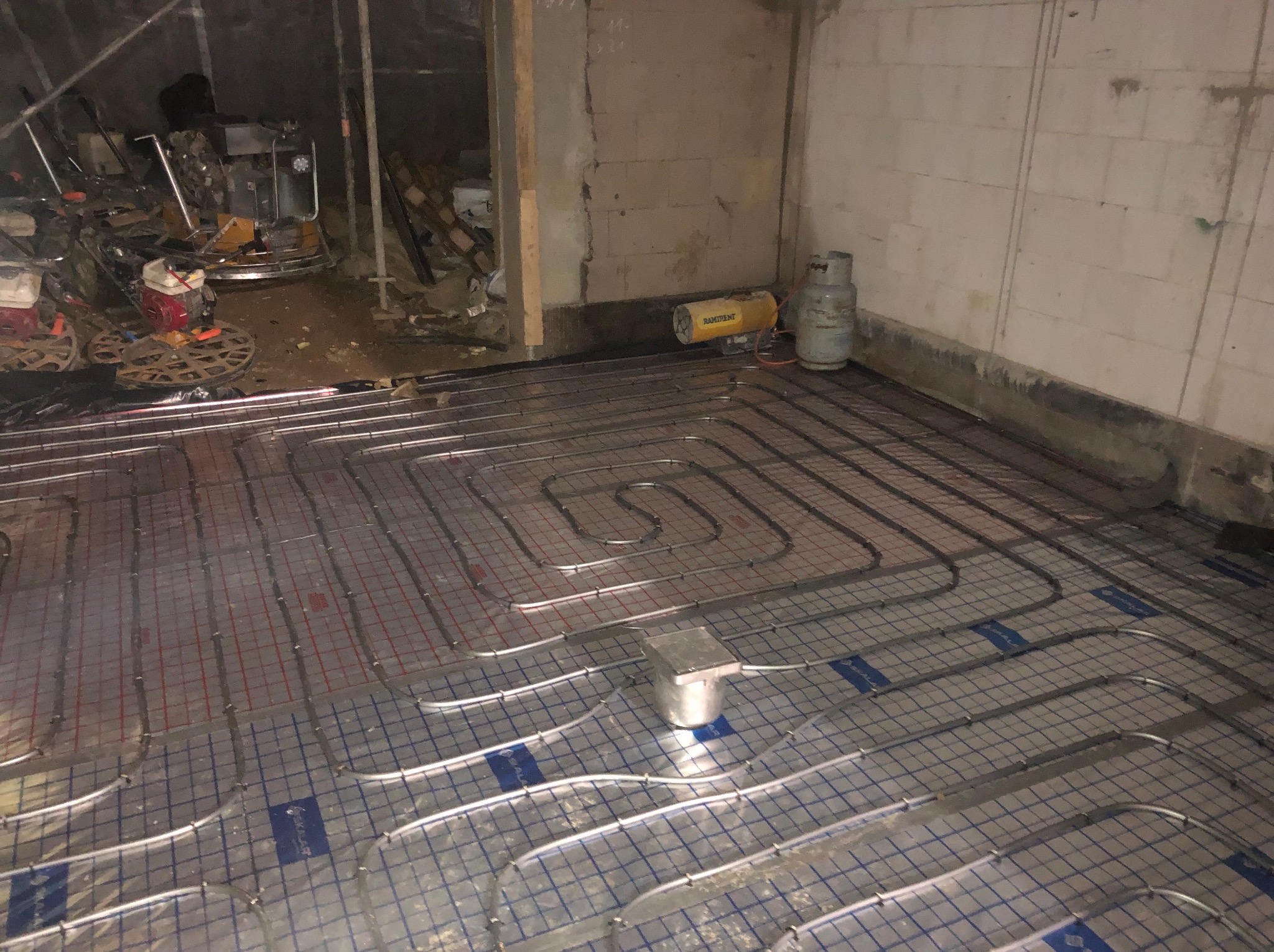 Ułożona instalacja ogrzewania podłogowego na folii izolacyjnej w surowym pomieszczeniu, widoczny nagrzewnica i butla gazowa w tle.