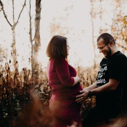 Inner Light - Fotografia Ślubna - Ciepłe ujęcie kobiety w bordowej sukience i mężczyzny obejmującego jej ciążowy brzuch na tle drzew w jesiennej aurze, delikatne światło, uśmiechnięte twarze.