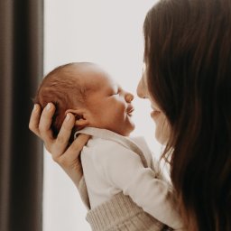 Inner Light - Fotografia Ślubna - Profil matki z długimi brązowymi włosami trzymającej noworodka w beżowym ubranku, delikatne światło, bliskość twarzy, neutralne tło.