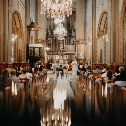 Inner Light - Fotografia Ślubna - Para młoda siedzi tyłem w kościele podczas ceremonii ślubnej. W tle ołtarz i ksiądz. W ławkach siedzą goście. Widoczne dekoracje kwiatowe i świece.