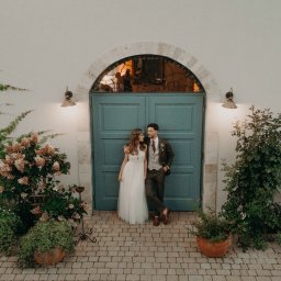 Inner Light - Fotografia Ślubna - Para młoda w stylizowanych strojach pozuje na tle błękitnych drzwi z kamiennym łukiem i roślinami w donicach. Ujęcie z góry, ciepłe oświetlenie.