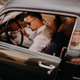 Inner Light - Fotografia Ślubna - Elegancki mężczyzna w okularach przeciwsłonecznych siedzi za kierownicą klasycznego, ciemnozielonego samochodu. Ubrany w białą koszulę i kamizelkę.