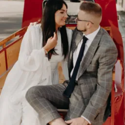 Uśmiechnięta para w stylizowanych strojach siedzi w czerwonym, zabytkowym samochodzie, spoglądając na siebie podczas sesji plenerowej.