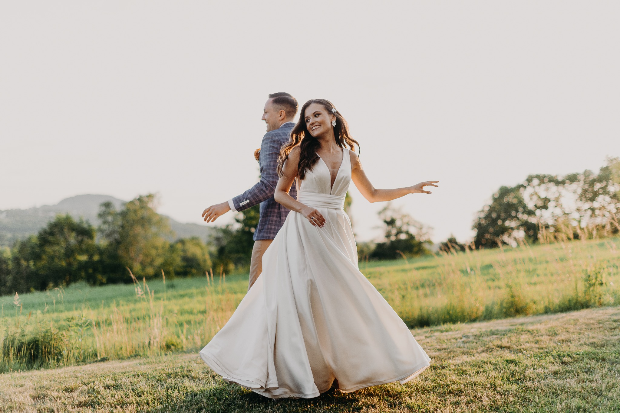 Para młoda stoi tyłem do siebie na łące. Panna młoda w białej sukni z rozcięciem uśmiecha się, a pan młody w niebieskiej marynarce patrzy w bok. Romantyczna sesja w plenerze.