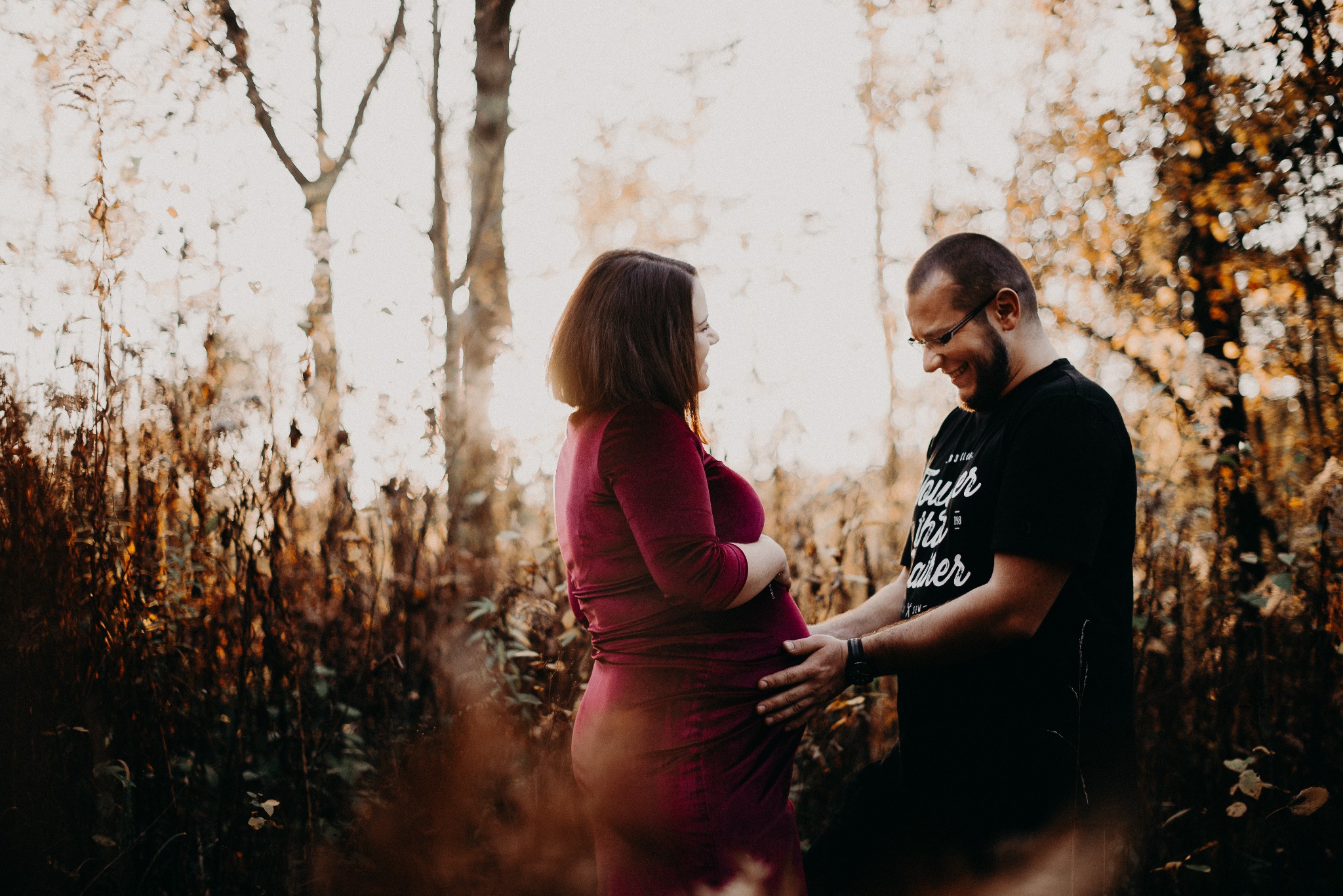 Ciepłe ujęcie kobiety w bordowej sukience i mężczyzny obejmującego jej ciążowy brzuch na tle drzew w jesiennej aurze, delikatne światło, uśmiechnięte twarze.