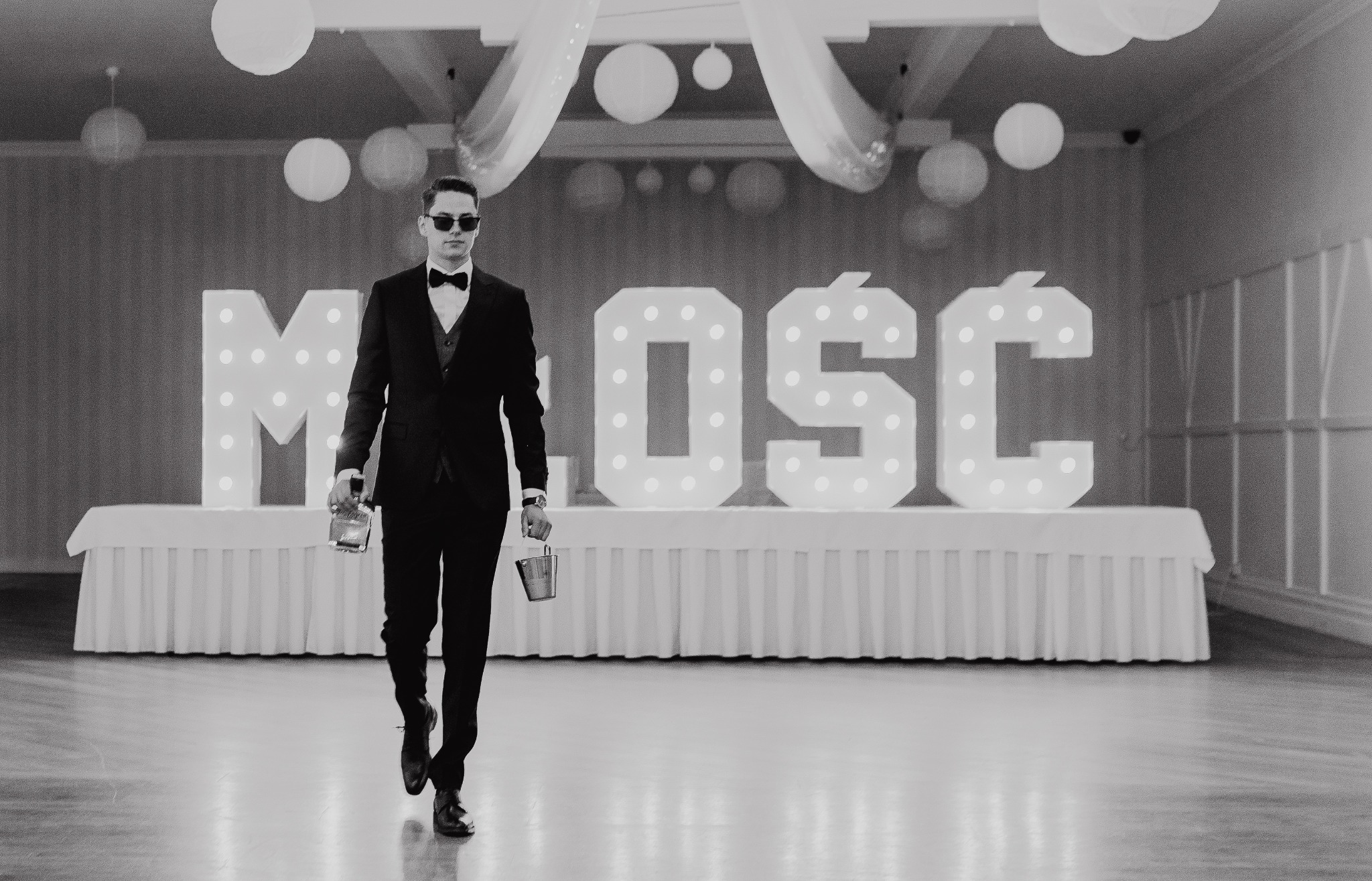 Czarno-białe zdjęcie mężczyzny w garniturze i okularach, idącego z wiadrami na tle napisu 'MIŁOŚĆ' z żarówkami, w sali udekorowanej lampionami.