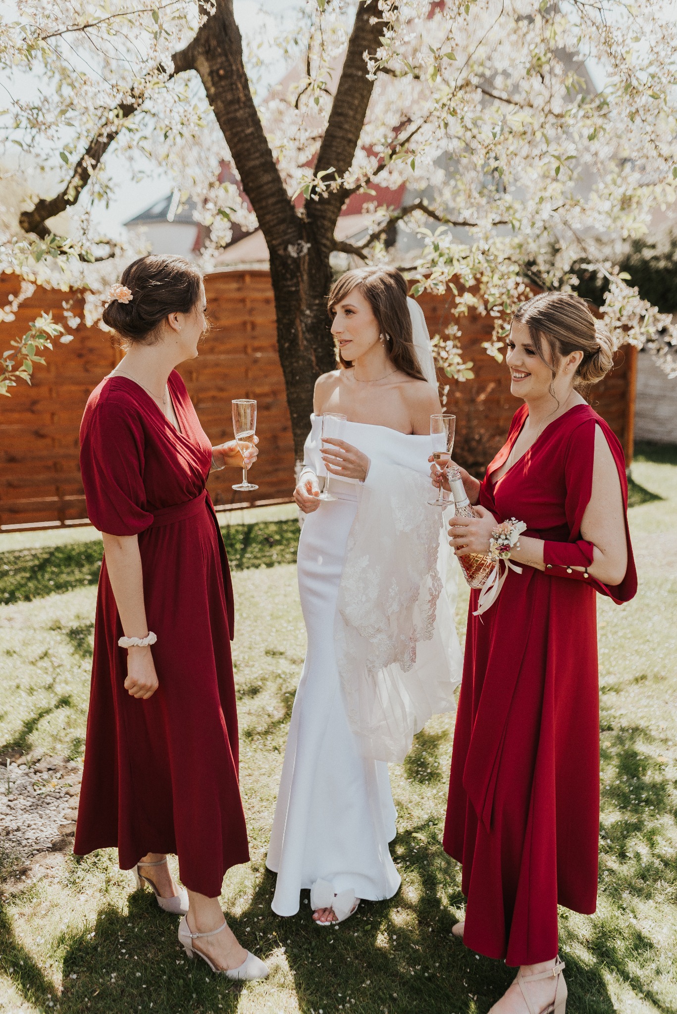 Panna młoda w białej sukni z druhnami w bordowych sukienkach, wznoszą toast szampanem w plenerze, w tle kwitnące drzewo.