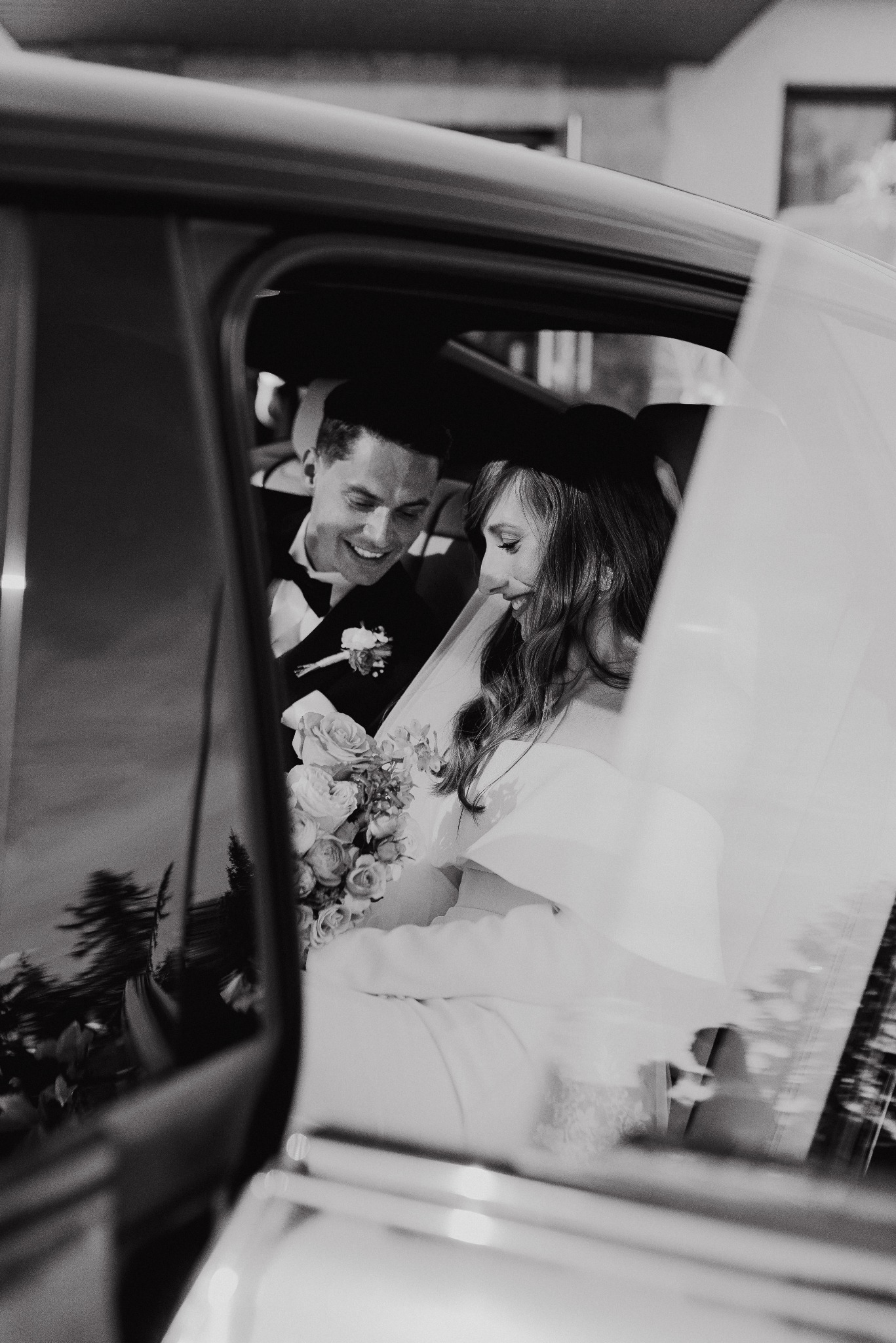 Czarno-białe zdjęcie nowożeńców w samochodzie: uśmiechnięty pan młody w smokingu i panna młoda z bukietem, spoglądająca w dół. Widok przez szybę, z delikatnym efektem rozmycia.