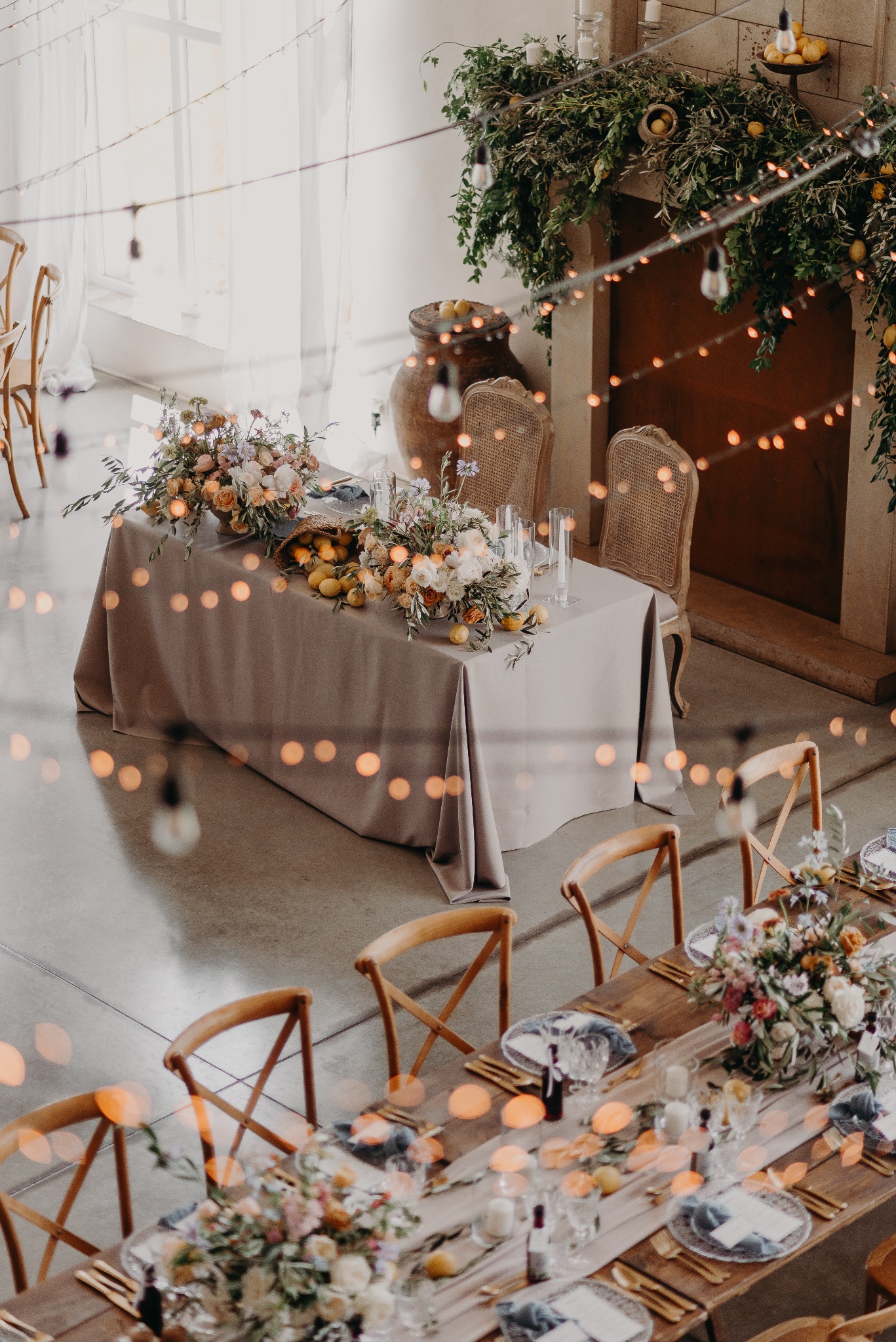 Udekorowany stół weselny z kwiatami, owocami i girlandami świetlnymi, widok z góry. W tle kamienny kominek z zieloną dekoracją i cytrynami. Elegancki wystrój.