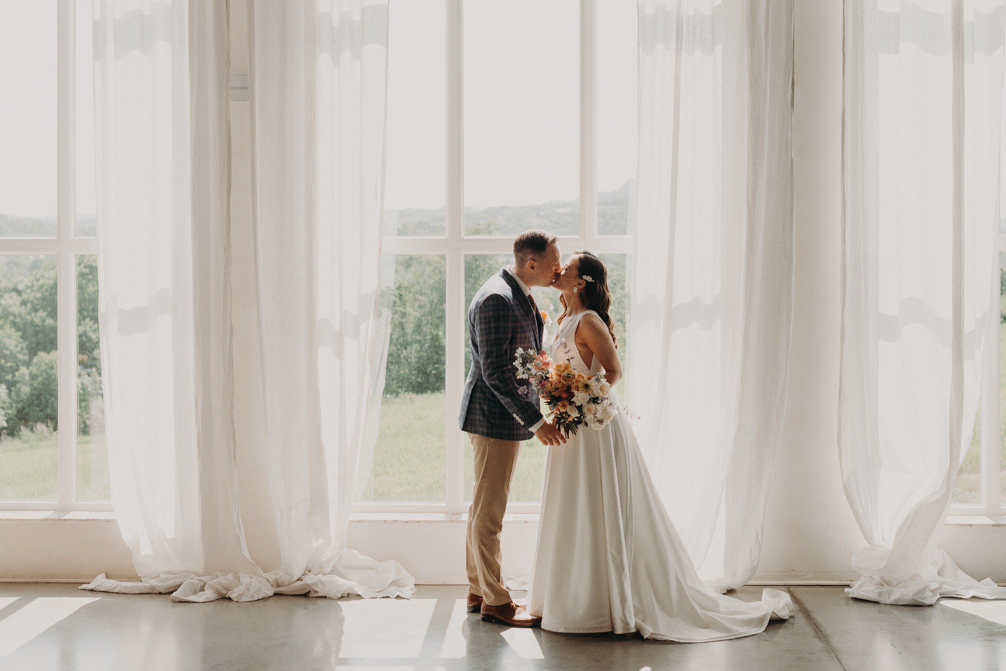 Para młoda w objęciach, delikatny pocałunek na tle okna z białymi zasłonami. Panna młoda trzyma bukiet. Subtelne światło podkreśla intymną chwilę.
