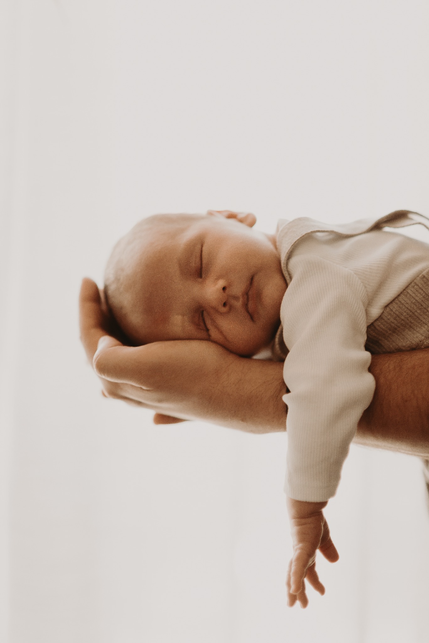 Delikatne ujęcie śpiącego noworodka, trzymanego w dłoniach dorosłego. Jasna tonacja, miękkie światło podkreśla niewinność dziecka.