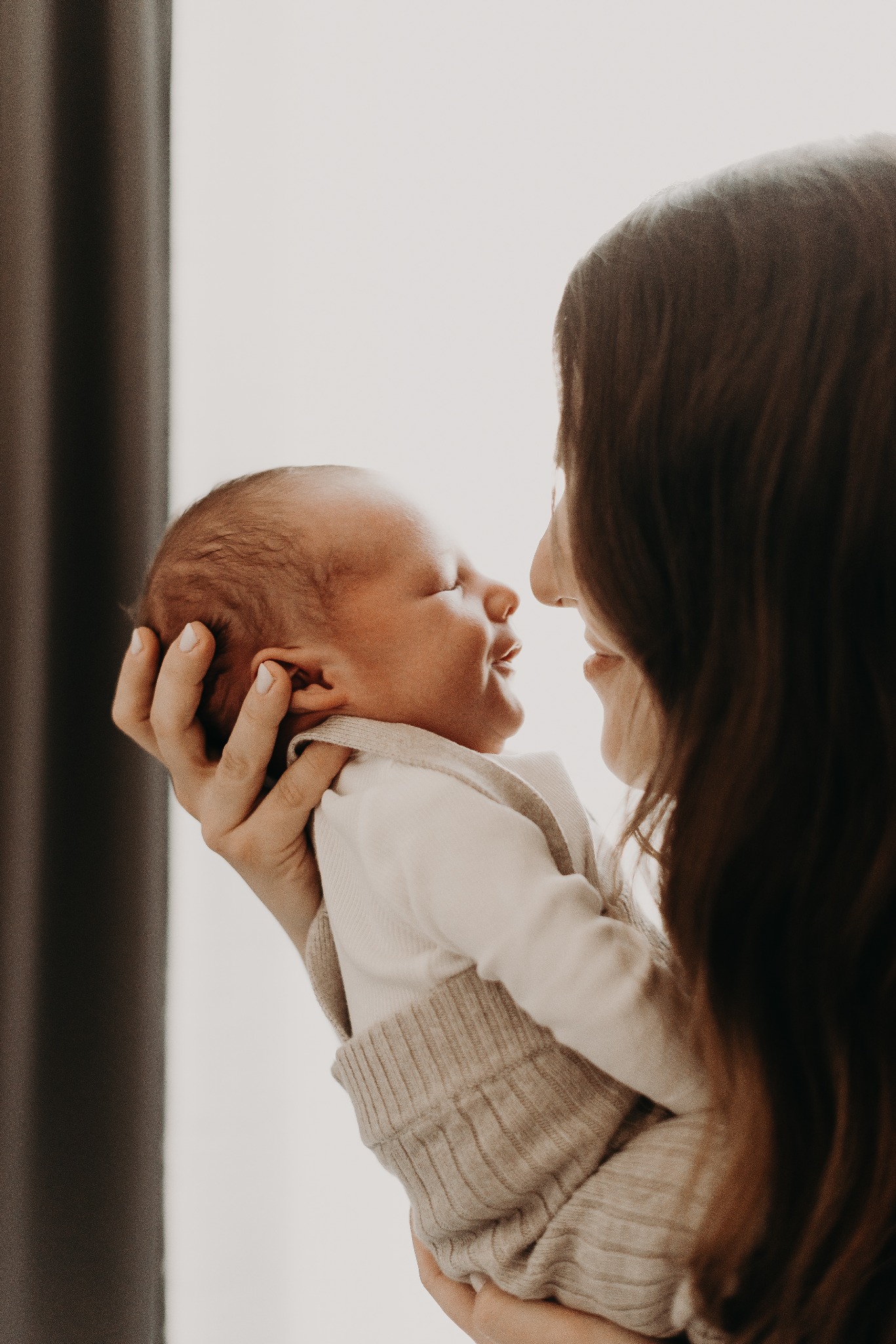 Profil matki z długimi brązowymi włosami trzymającej noworodka w beżowym ubranku, delikatne światło, bliskość twarzy, neutralne tło.