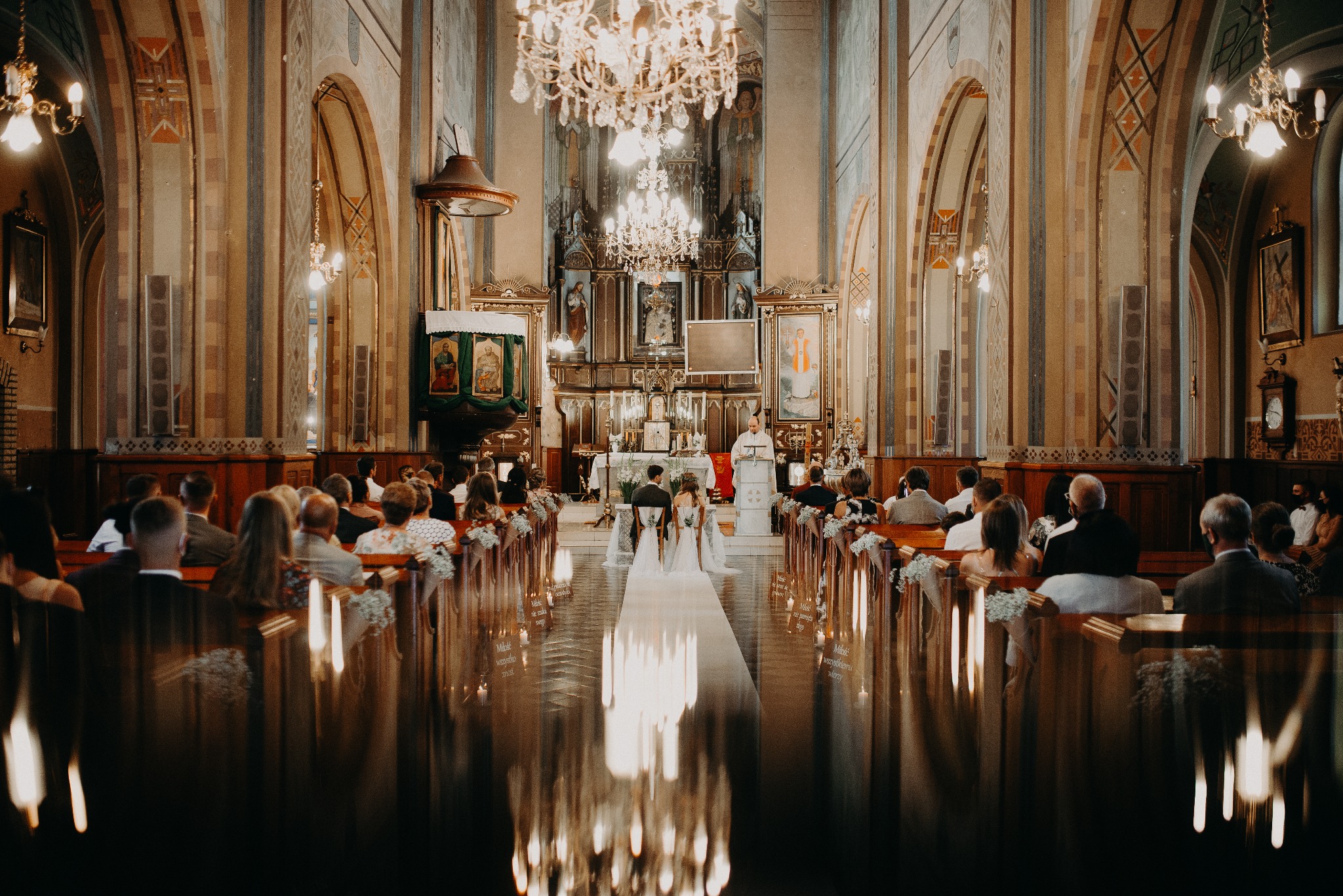 Para młoda siedzi tyłem w kościele podczas ceremonii ślubnej. W tle ołtarz i ksiądz. W ławkach siedzą goście. Widoczne dekoracje kwiatowe i świece.